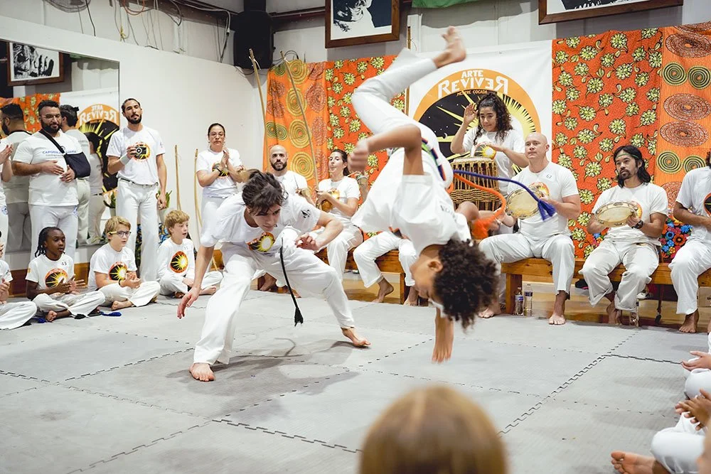 Two Capoeira practitioners performing a flip during a group demonstration, surrounded by spectators and musicians in a decorated indoor space.