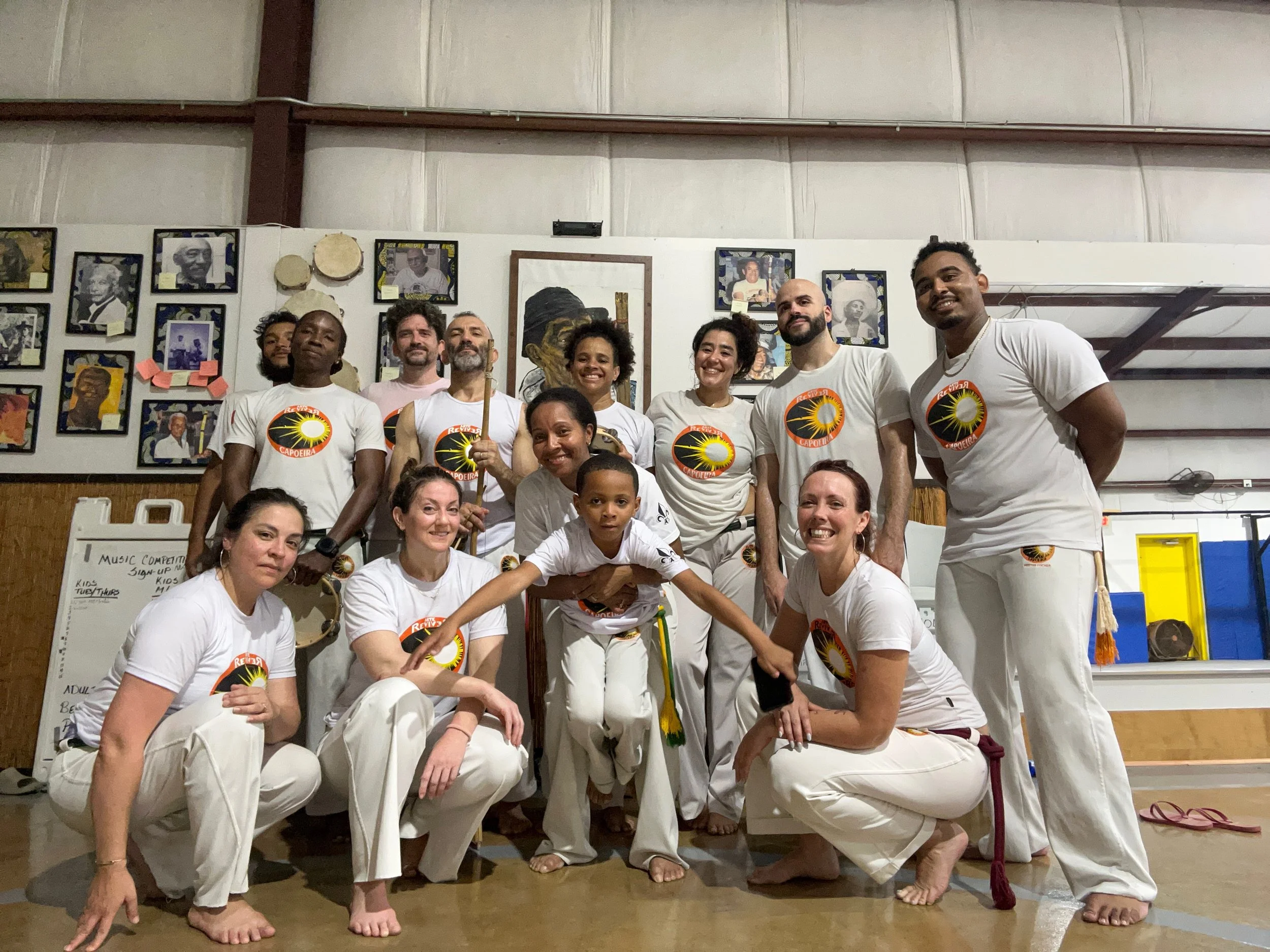 A group of people participating in a capoeira class or demonstration, dressed in white uniforms with a capoeira logo, posing together in an indoor setting with artwork and photos on the wall behind them.