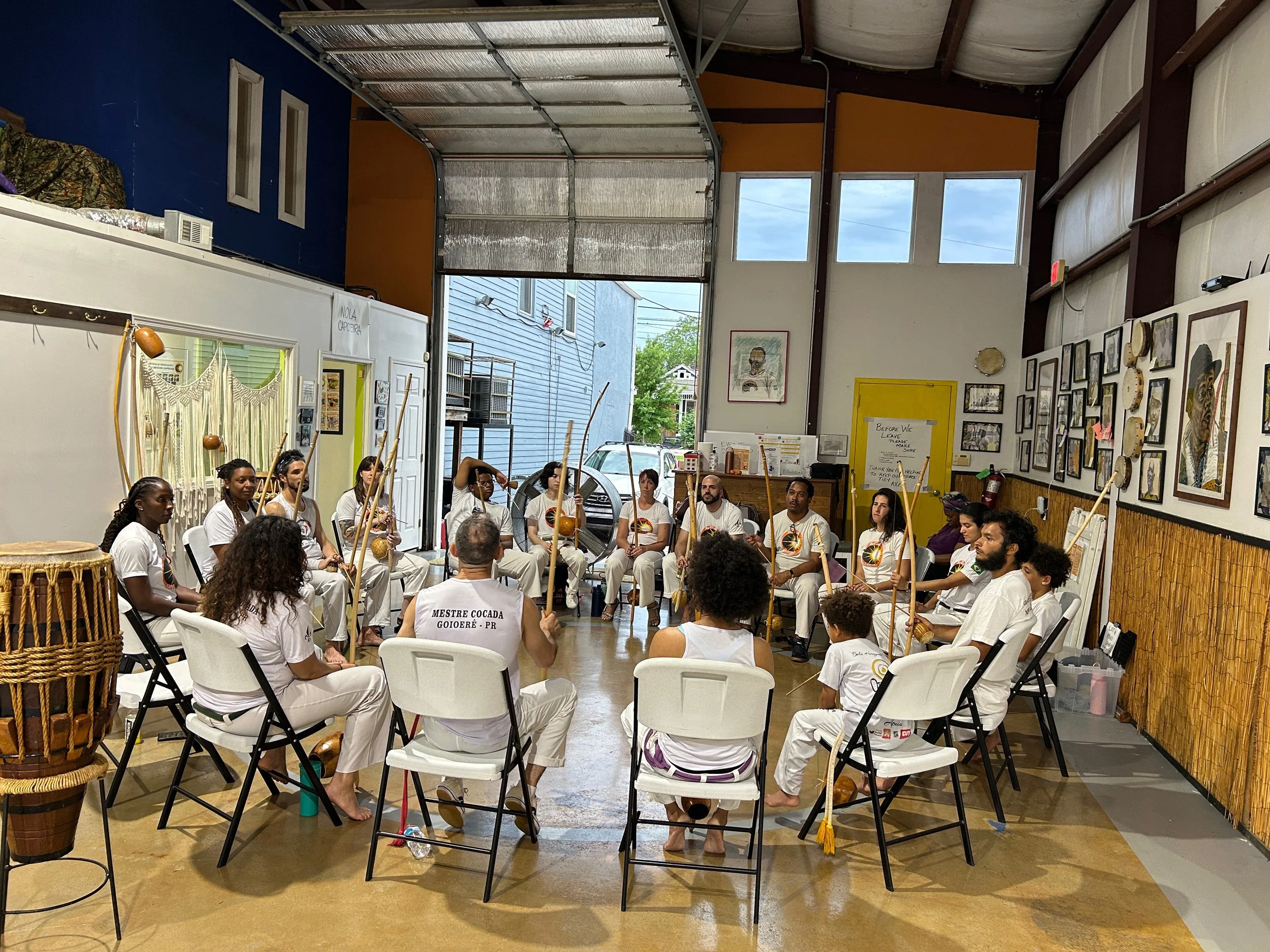 A group of people dressed in white, sitting on white chairs in a spacious indoor area with tall windows and art on the walls, participating in a cultural or training event involving the use of sticks and drums.