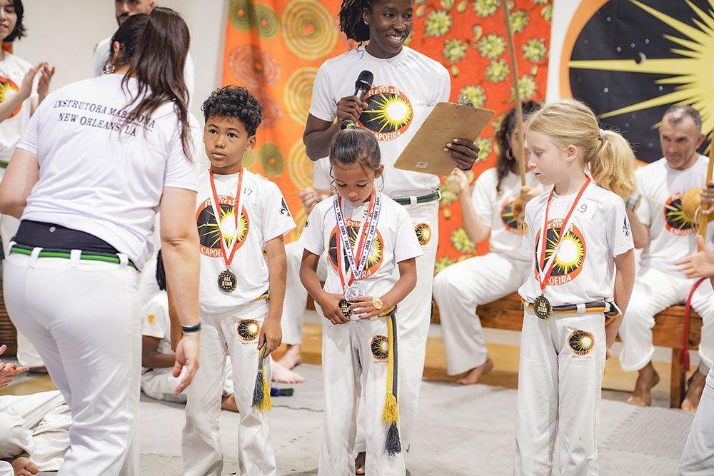 Children receiving awards at a Capoeira event, with medals around their necks, in a decorated room with a large clock and vibrant orange and yellow backdrop.