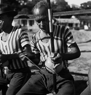 A black and white photograph of Mestre Pastinha playing berimbau while seated on a bench wearing his traditional striped uniform.