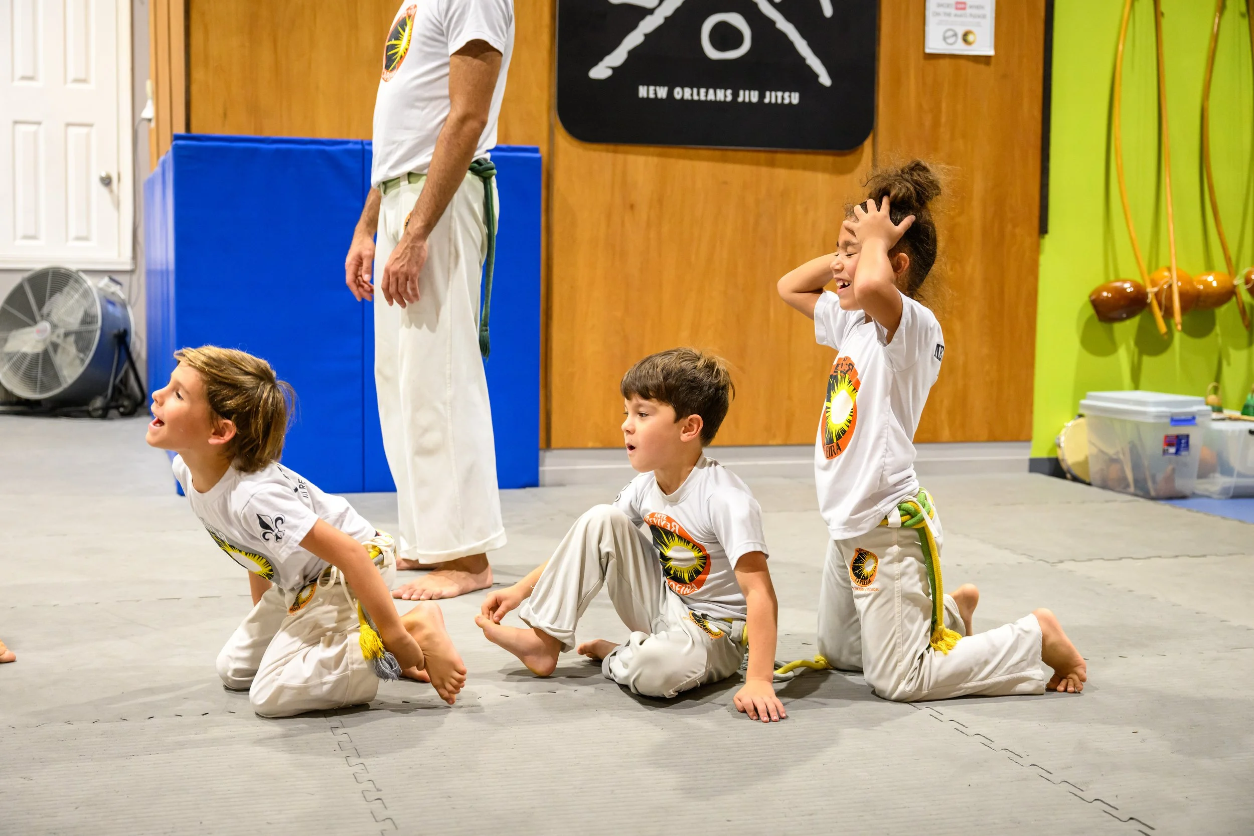 Young children in martial arts uniforms kneeling on the floor during a class, with a woman standing nearby, involved in a martial arts training session at a dojo.