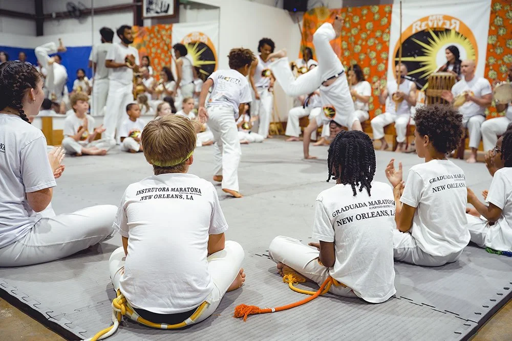 Children and adults practicing capoeira in a gym or community center, with some children seated on the floor and others performing acrobatic movements, wearing white uniforms with 