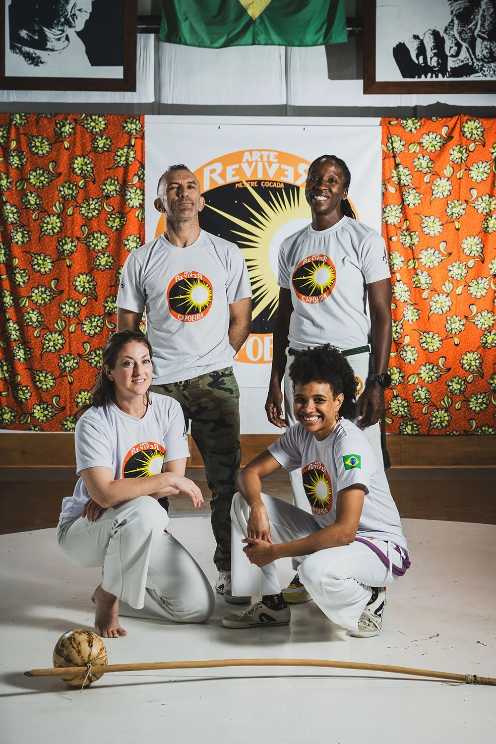 Group of four diverse people wearing white t-shirts with a sun logo, posing in front of colorful patterned curtains and a banner that reads 'Arte Revives Me Trec Cocado' with a sun and black rays. A traditional wooden bow and a gourd resting on the f