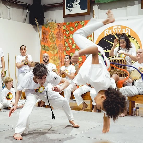 People practicing capoeira, a Brazilian martial art, indoors with some holding musical instruments, colorful curtains, and a large Revive Artes logo in the background.