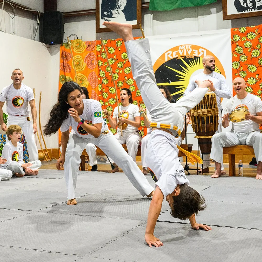 Two martial artists performing a Capoeira move during a group practice or performance, with other participants and musicians seated and standing around them in a decorated indoor space.