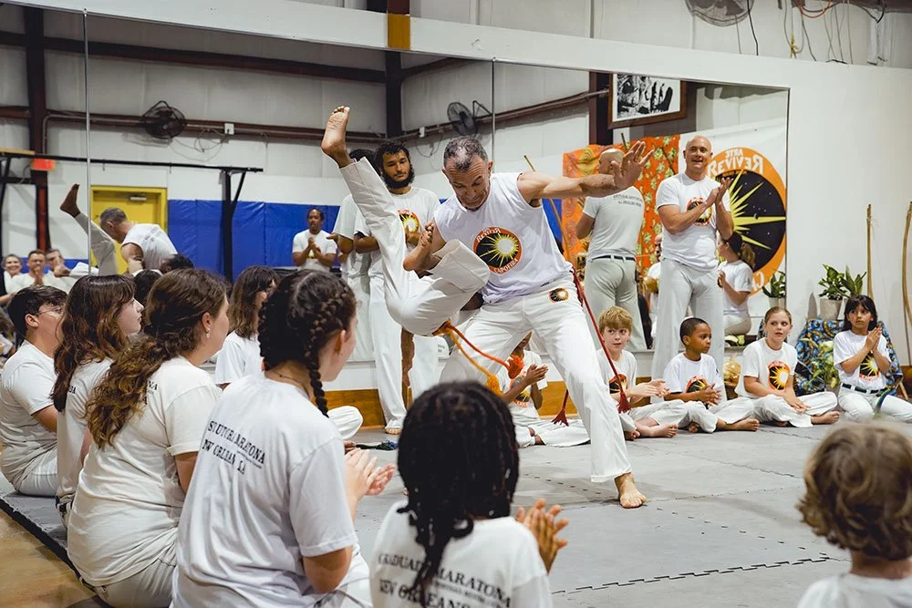 A martial arts class in a gym, with an instructor demonstrating a move to children and adults. Students are seated on the floor, watching the instructor perform a high kick. The gym has martial arts banners and equipment in the background.