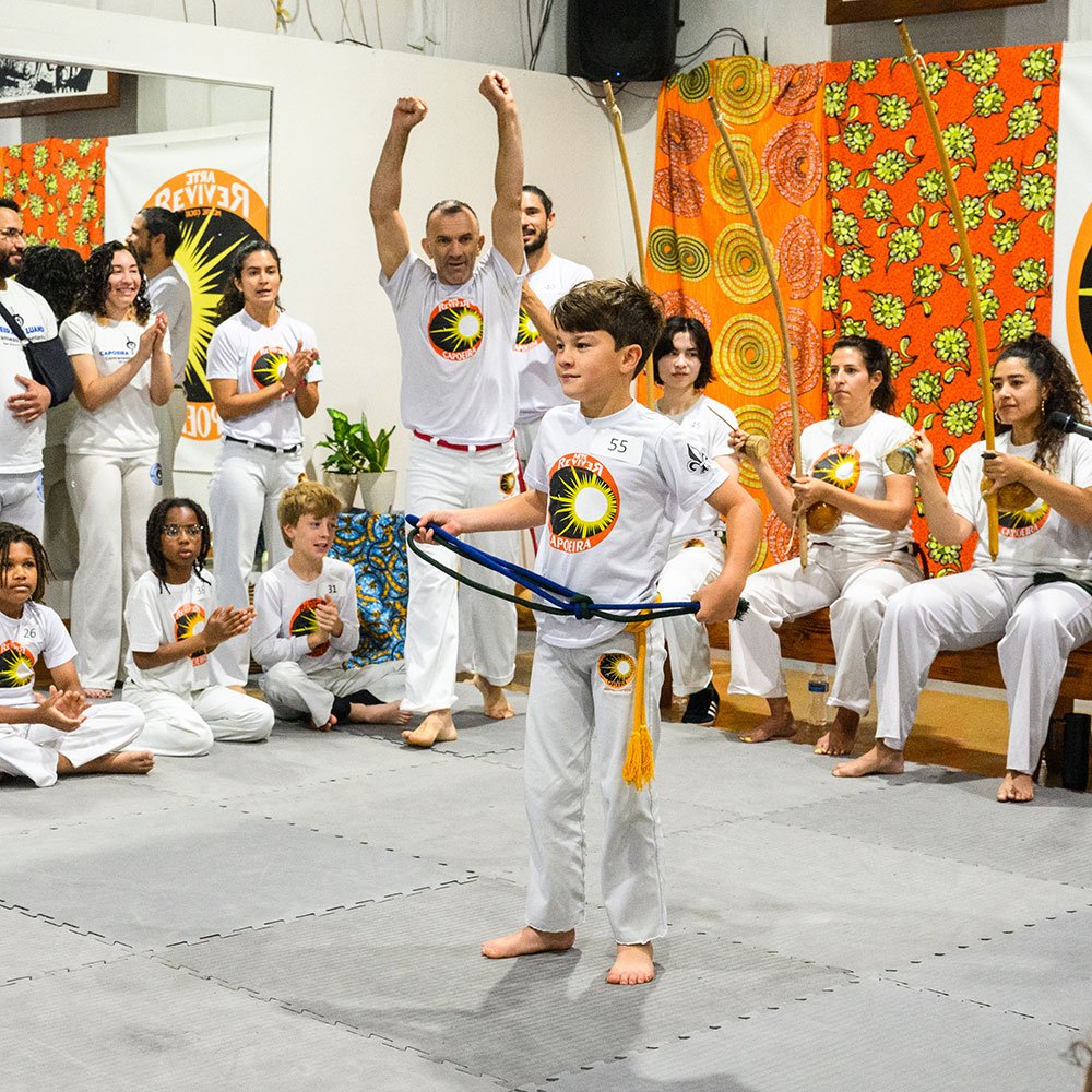 A boy practicing capoeira with a rope in a room decorated with colorful curtains, while group of children and adults clap, watch and perform capoeira moves, some holding traditional instruments, and a man raises his arms celebrating.