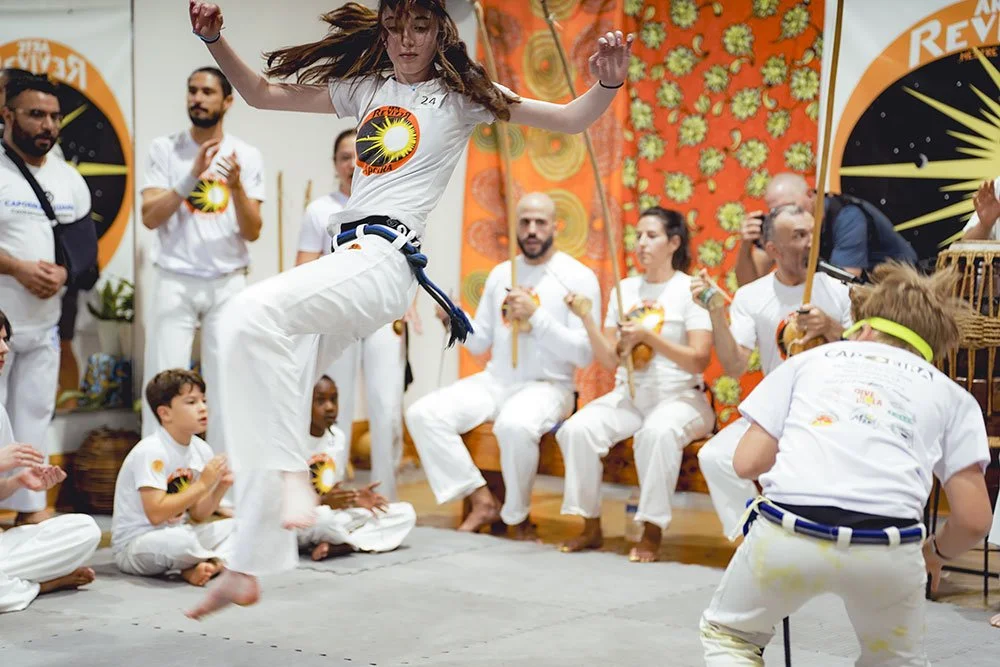 Group of people practicing Capoeira, a Brazilian martial art, in a room with vibrant orange floral-patterned walls and a large clock, some participants are sitting, others are standing, with a woman jumping in the center.