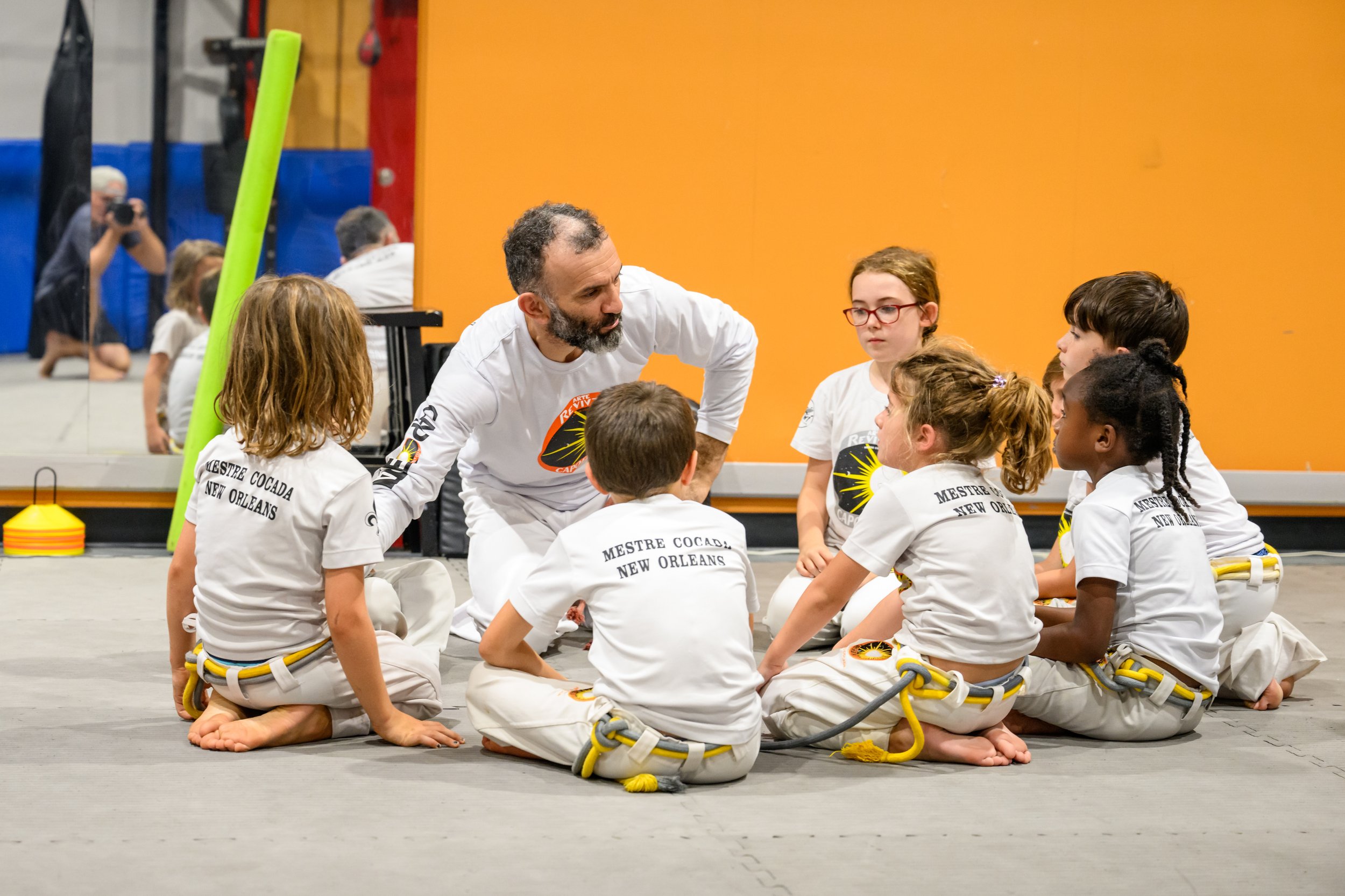 A coach speaking to a group of children sitting cross-legged on a gym floor during a martial arts class at Mestre Cocada in New Orleans. The children are wearing white martial arts uniforms with yellow belts and are attentively listening. The backgro