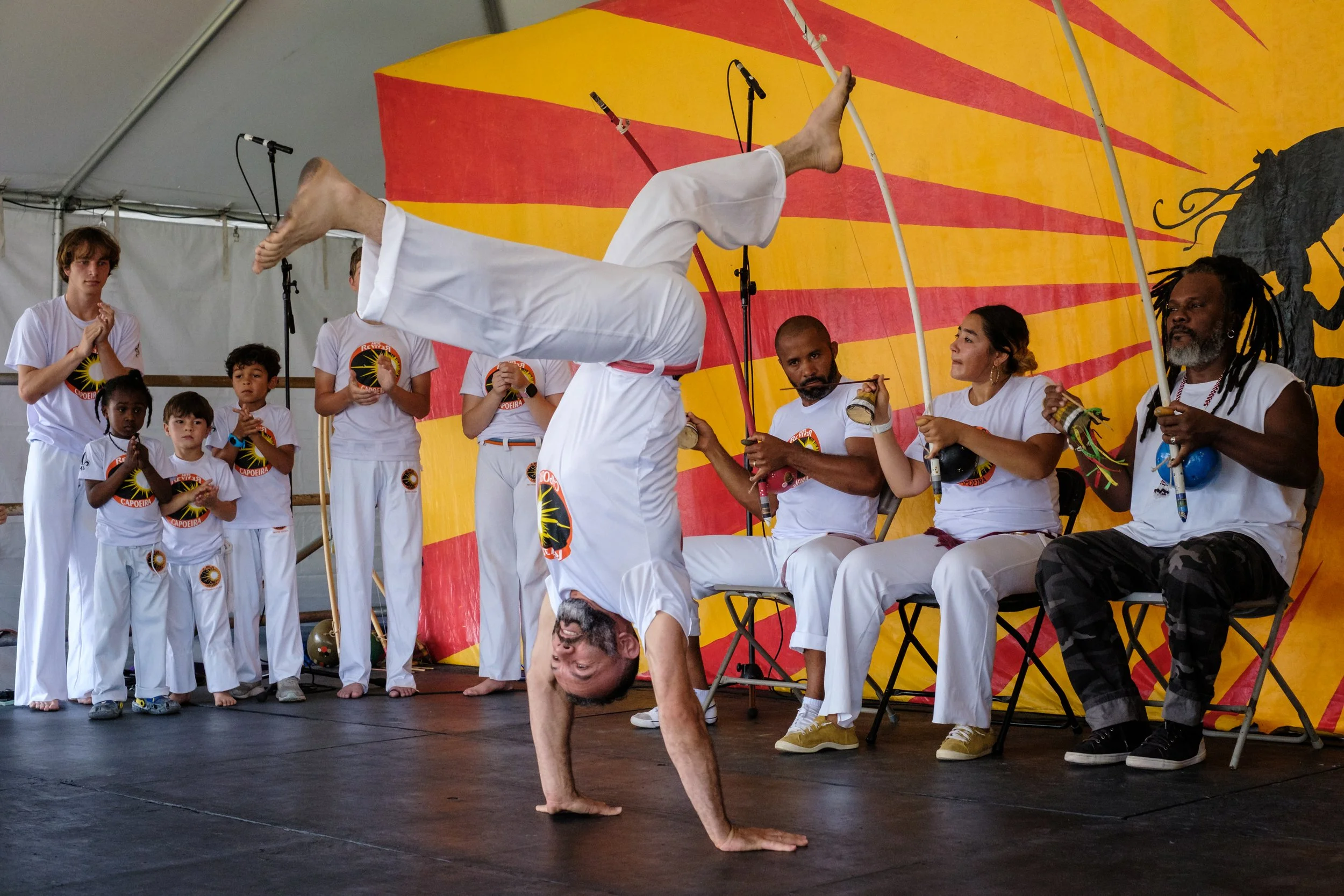 A man performing a handstand on a stage during a capoeira demonstration, with children and adults watching, some clapping and others holding musical instruments, in front of a colorful backdrop featuring a black silhouette of a lion.
