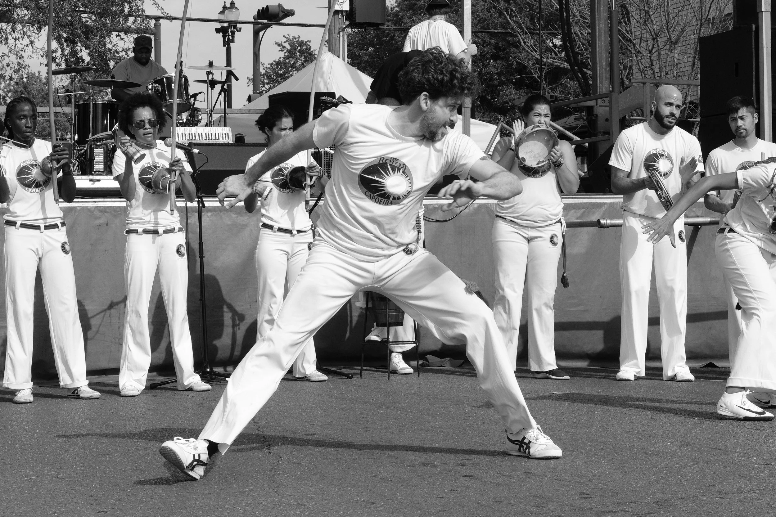 A group of musicians and dancers performing outdoors, with some holding percussion instruments and a man in the foreground dancing energetically.