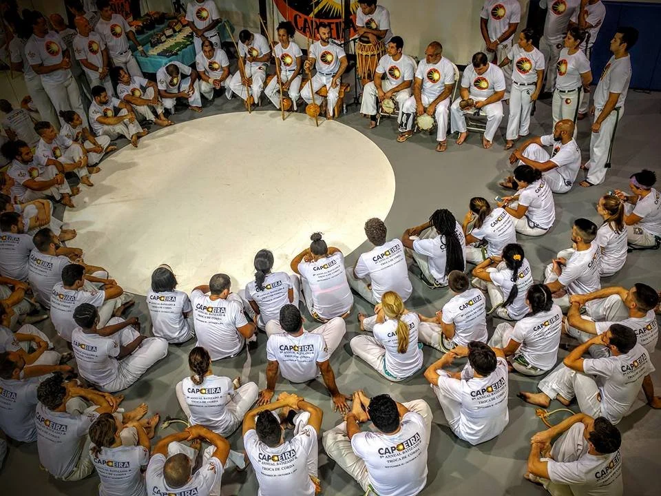 Group of people wearing white shirts and pants sitting and standing in a circle around a white circular space, participating in capoeira, a Brazilian martial art that combines dance, acrobatics, and music.
