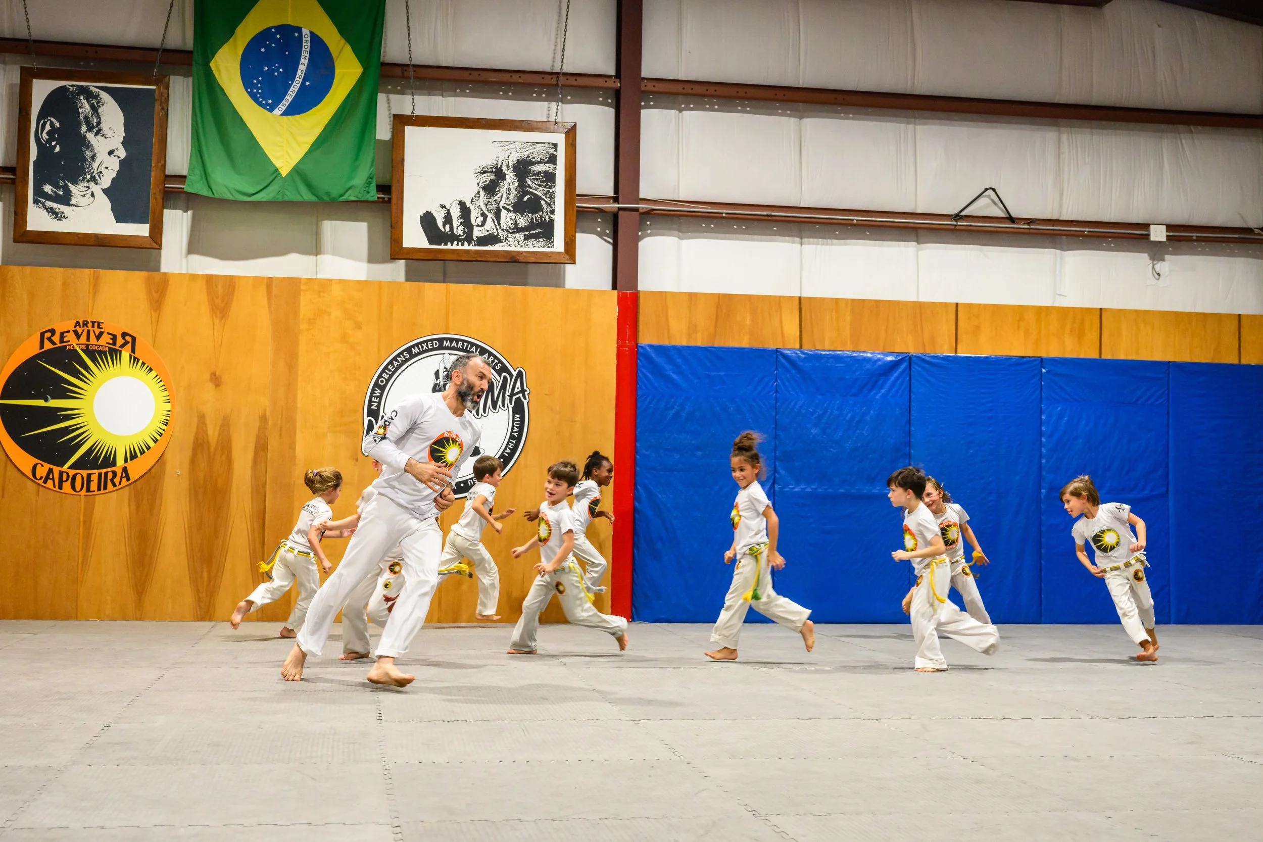A group of children in white martial arts uniforms with yellow belts and a man in similar attire practicing capoeira in a gymnasium, with Brazilian flags and portraits on the wall behind them.