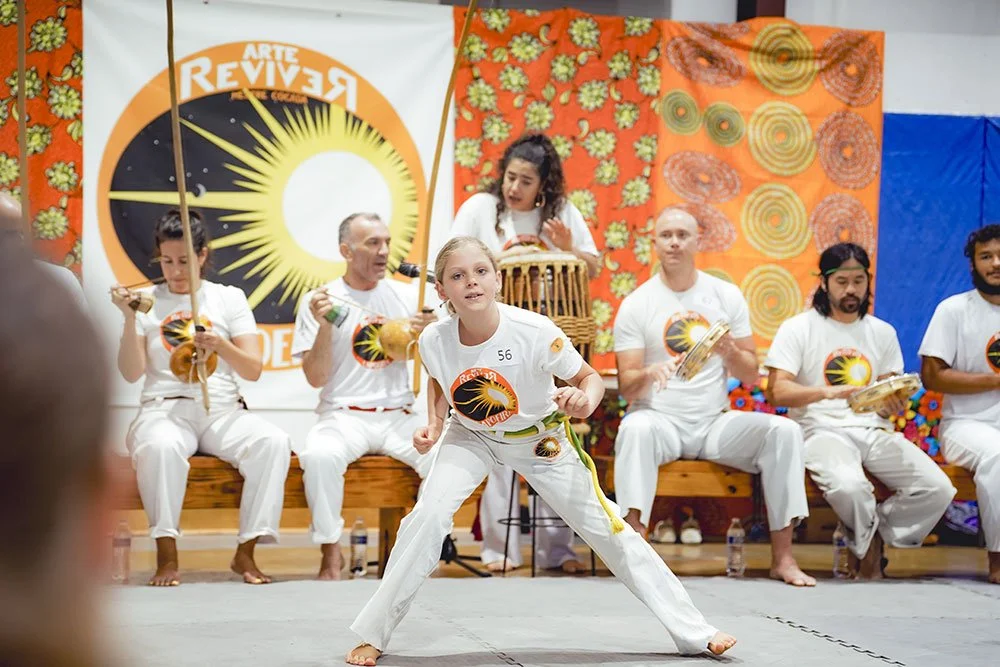 A young girl performing a martial arts move in front of a group of people playing traditional Brazilian instruments, with colorful decorations and a banner in the background.