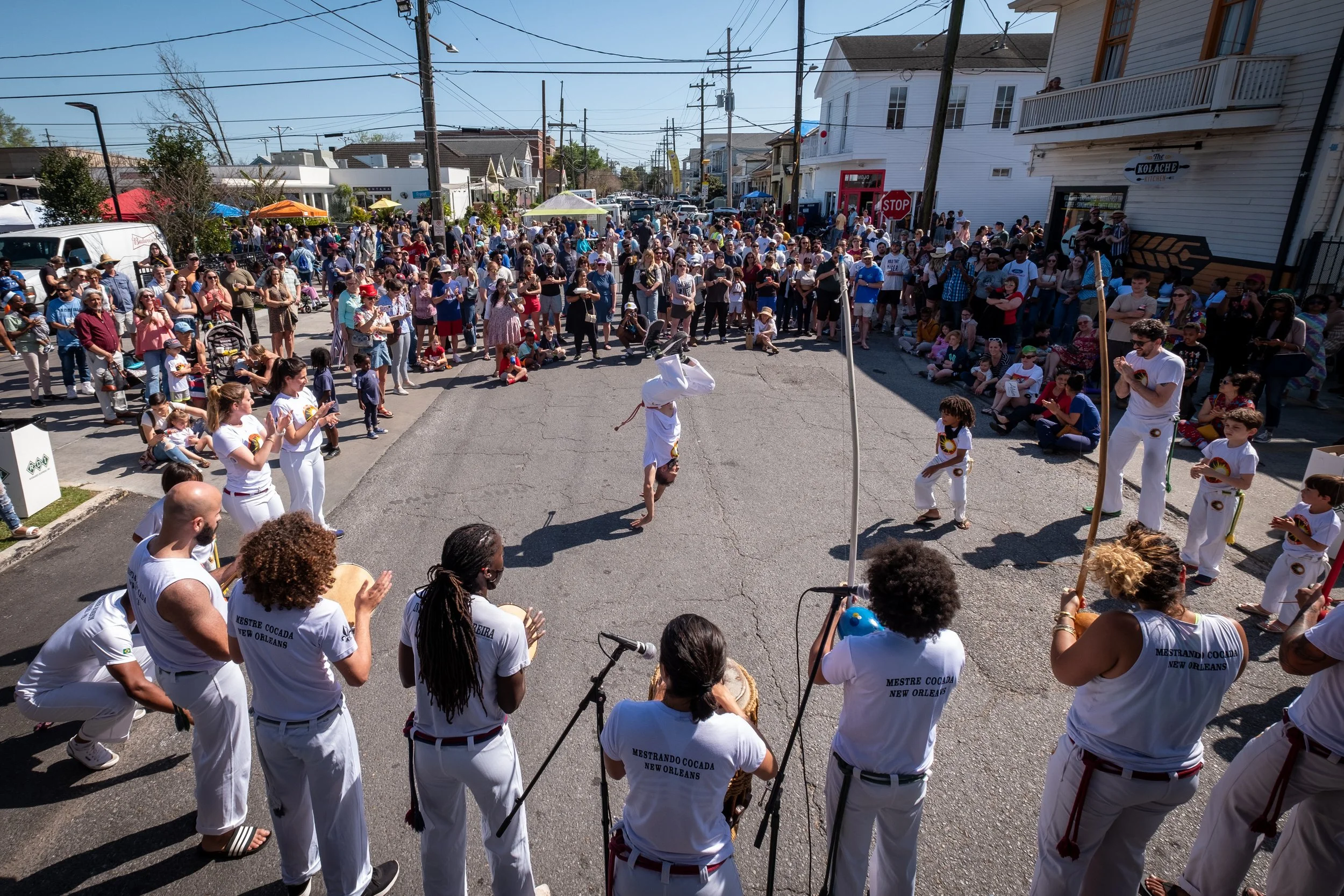 Street scene during a parade or public event with many people gathered. Performers in white uniforms are in the foreground, one executing a handstand. Audience members are sitting and standing along the street, some taking photos. The background feat