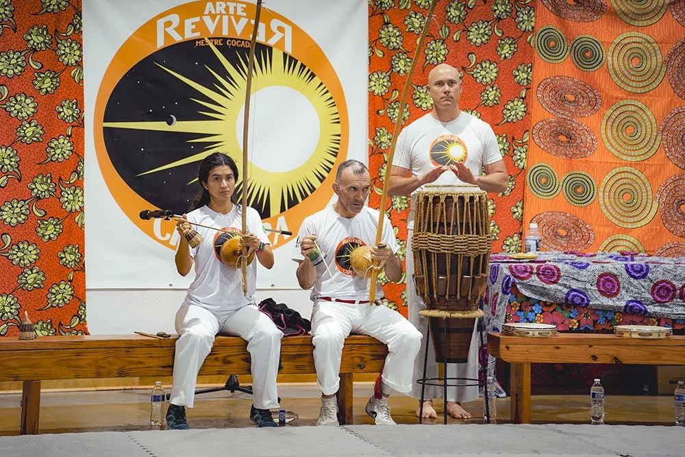 Three people dressed in white are seated or standing in front of a colorful backdrop with a logo for Arte Revira. They are holding musical instruments made from gourds with strings and sticks, and there is a large traditional drum on a stand. The sce