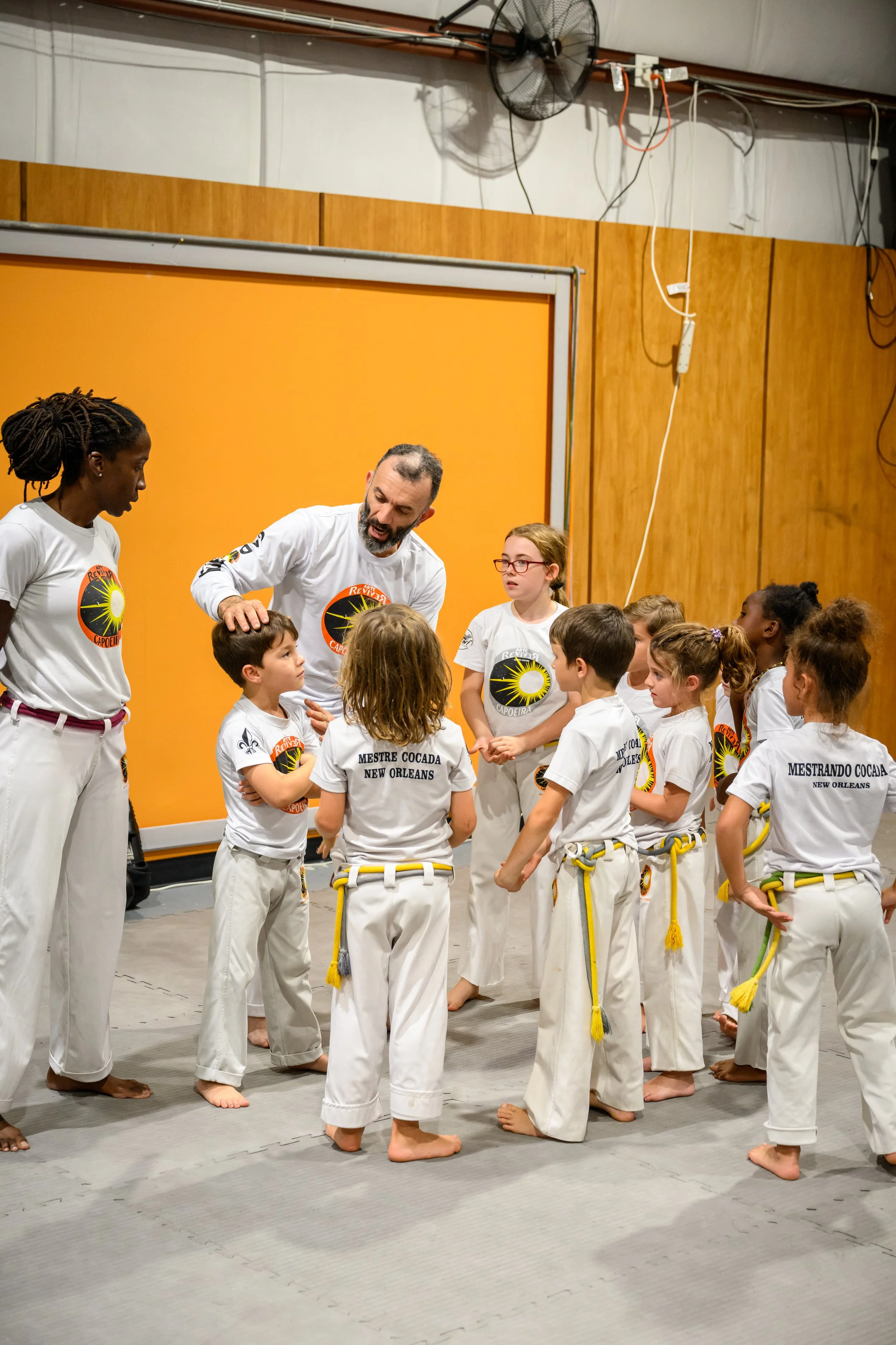 Group of children and adults wearing martial arts uniforms, standing on a dojo floor, listening to an instructor.