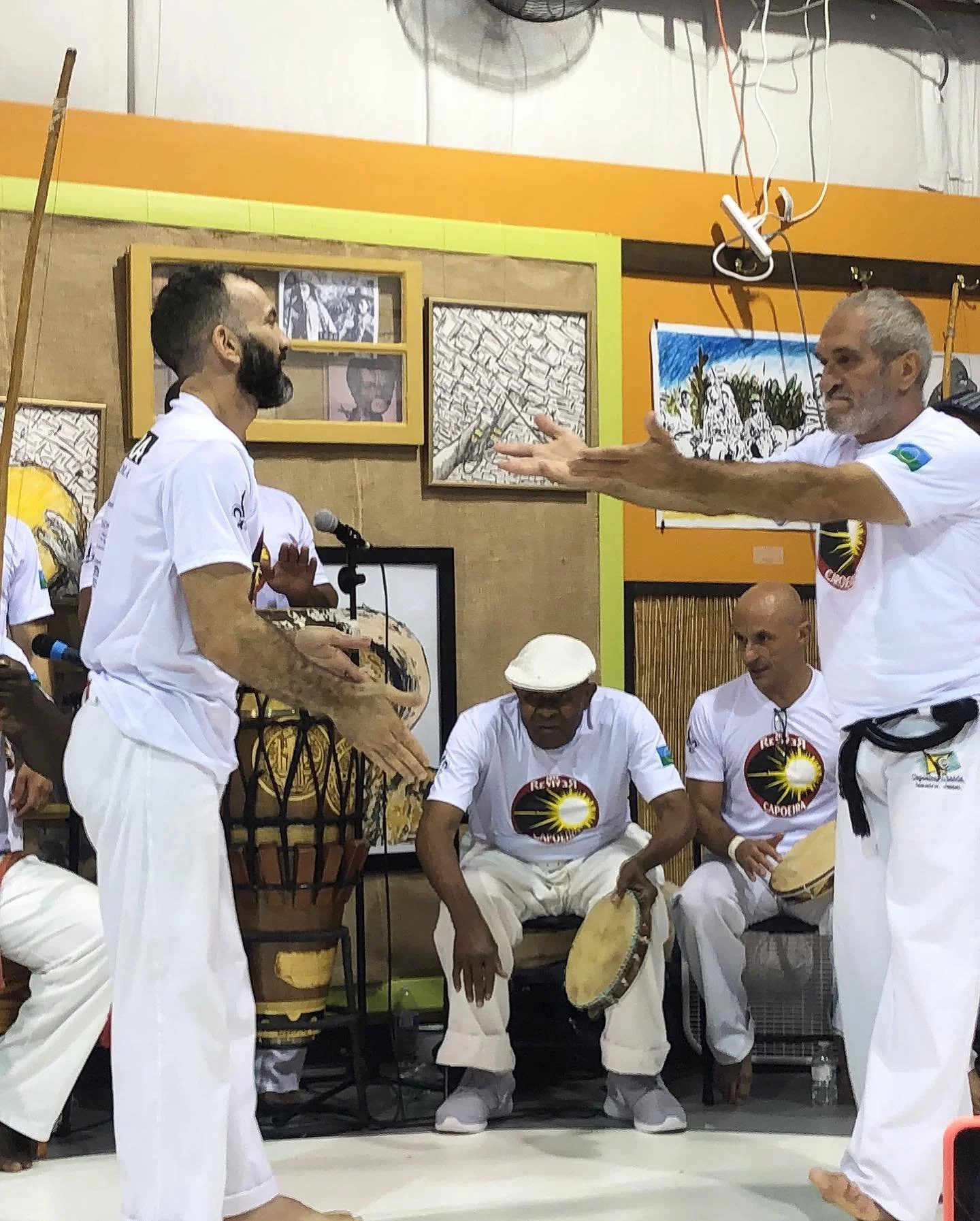 Group of people in white clothing playing traditional drums and percussion instruments in a room with artwork on the walls.