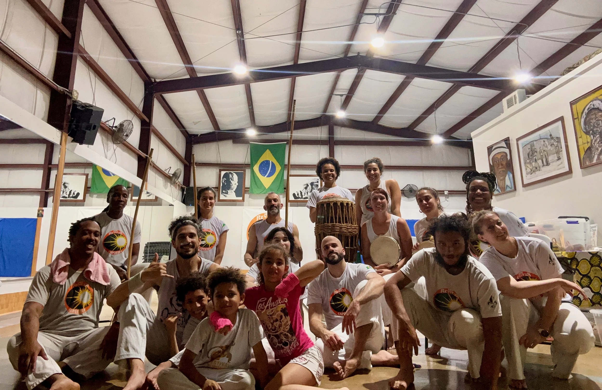 Group of people gathered in an indoor space with Brazilian flags hanging on the wall and artwork. Some individuals are holding drums, and many are dressed in white tank tops with a circular logo. The group appears to be celebrating or participating i