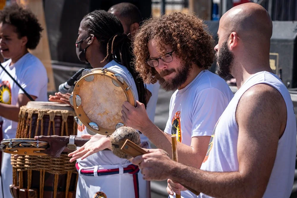Group of people playing various percussion instruments outdoors, including tambourine and drums.