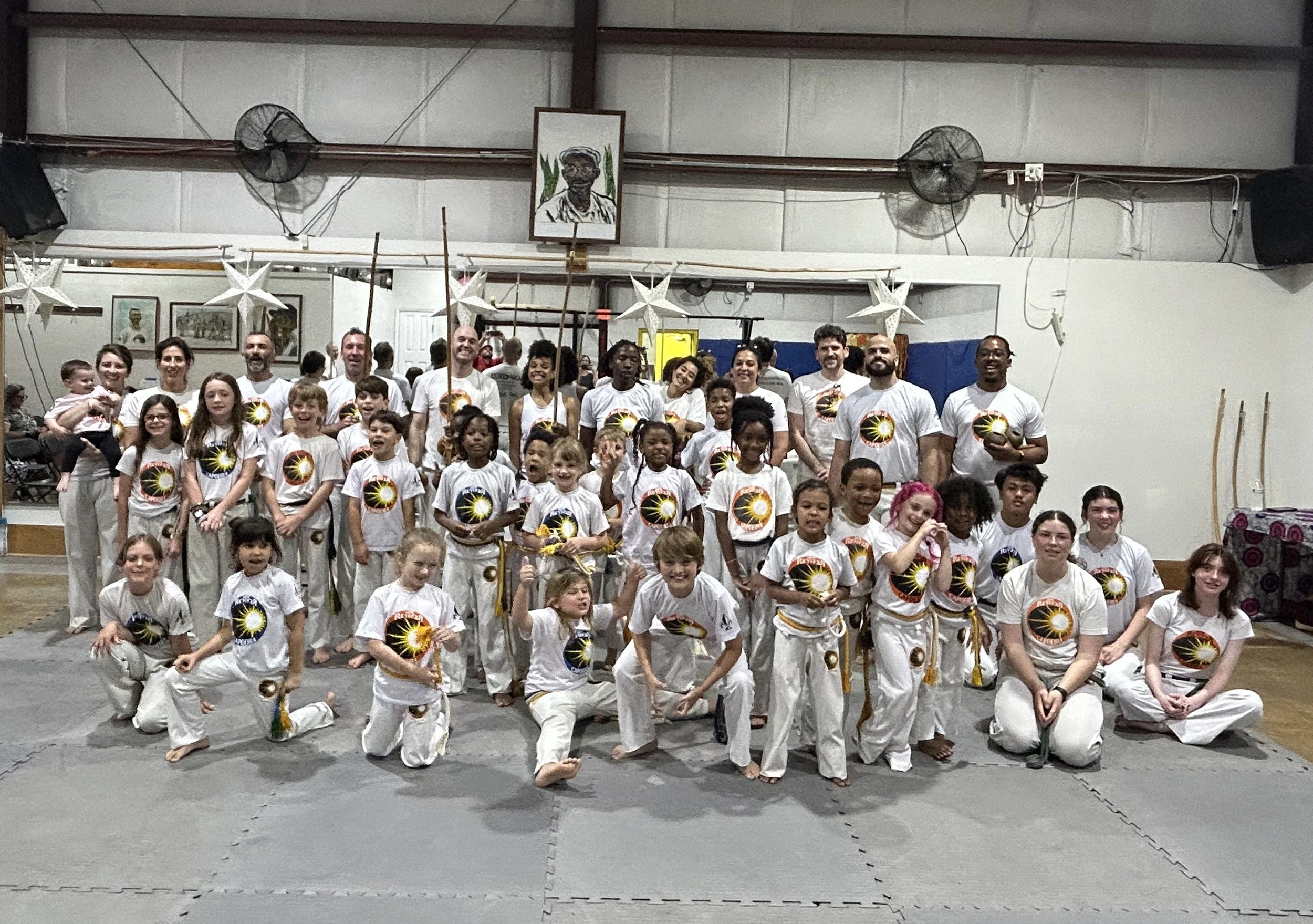 Group of people, including children and adults, in martial arts uniform and belts, inside a gym or martial arts dojo, posing for a photo with some holding martial arts weapons. Decorations include stars hanging from the ceiling, and a portrait of a p