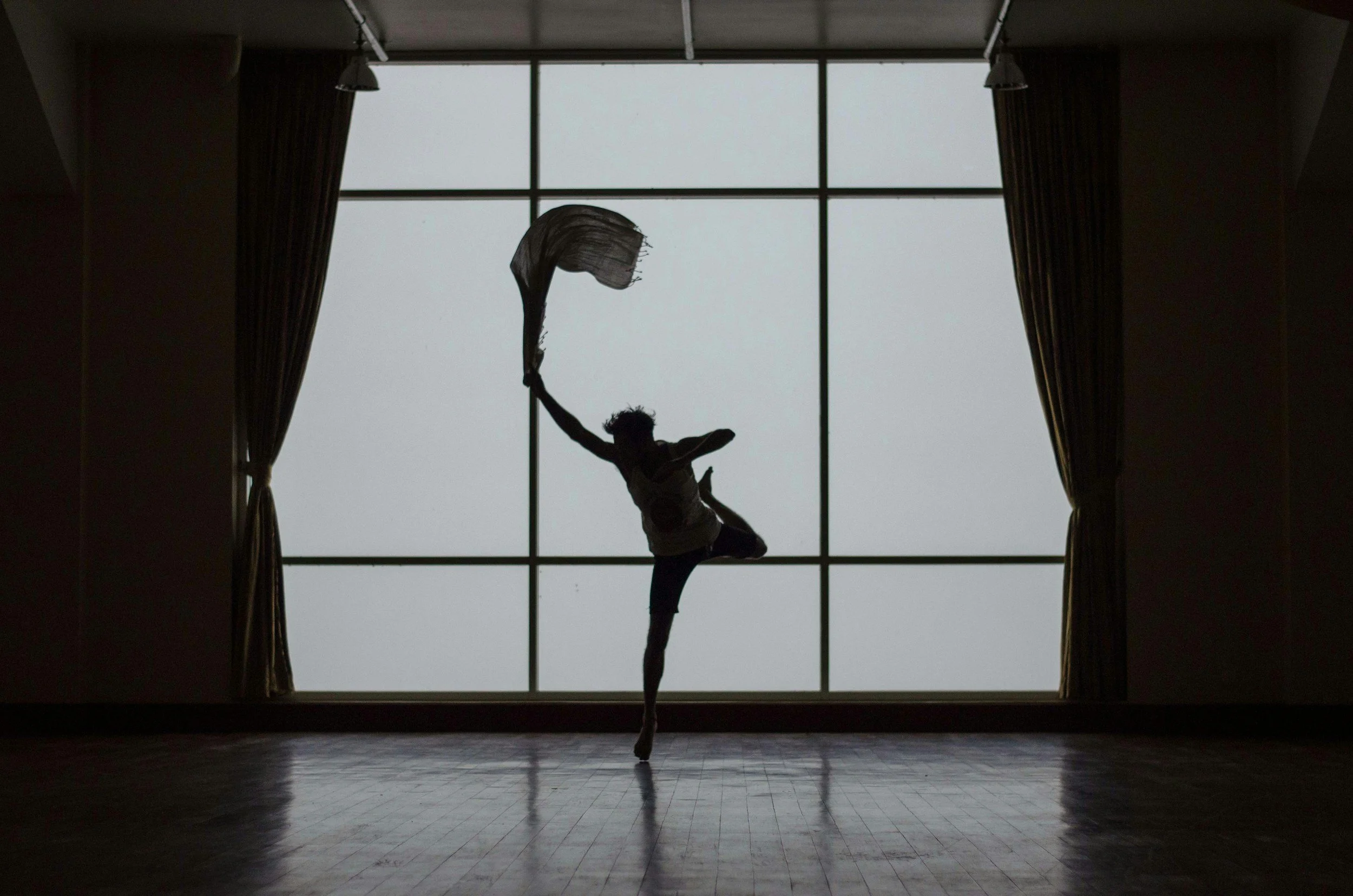 A silhouette of a person performing a dance or gymnastics move in front of a large window with curtains on each side.