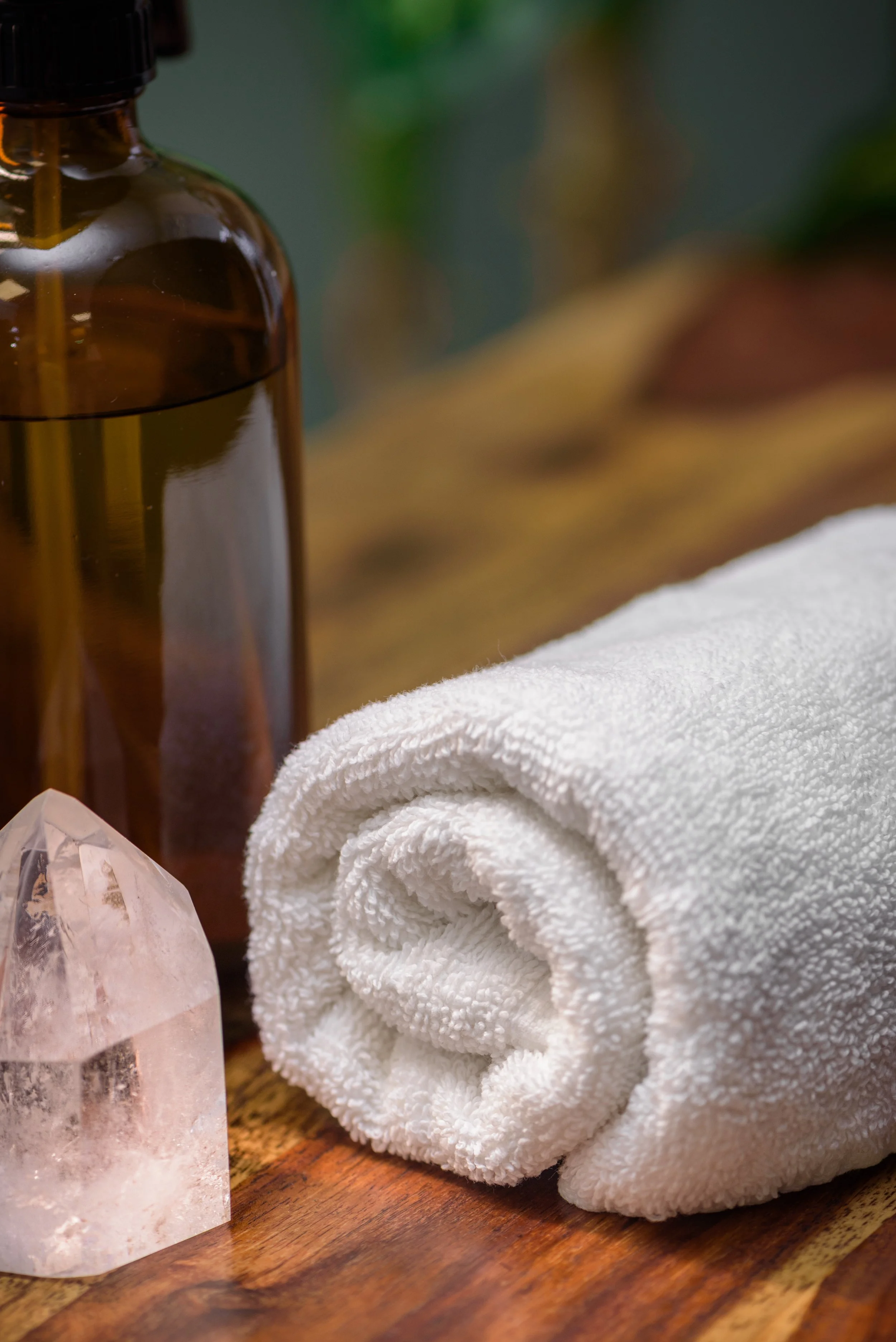 A brown glass bottle, white rolled towel, a large pink Himalayan salt crystal, and part of a patterned fabric on a wooden surface, with a blurred green and beige background.