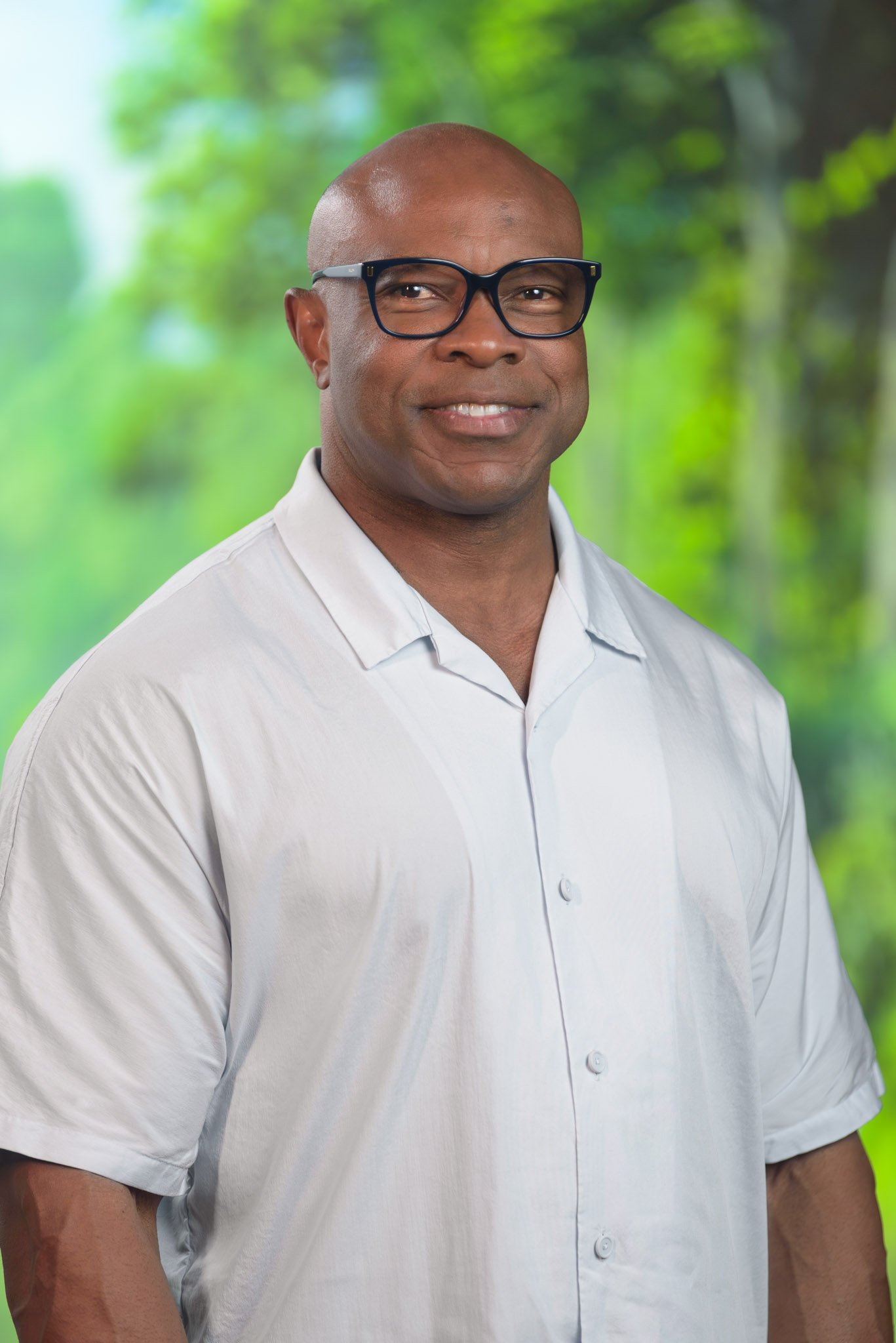 Headshot of a man with glasses, wearing a white shirt, standing outdoors with green blurred trees in the background.