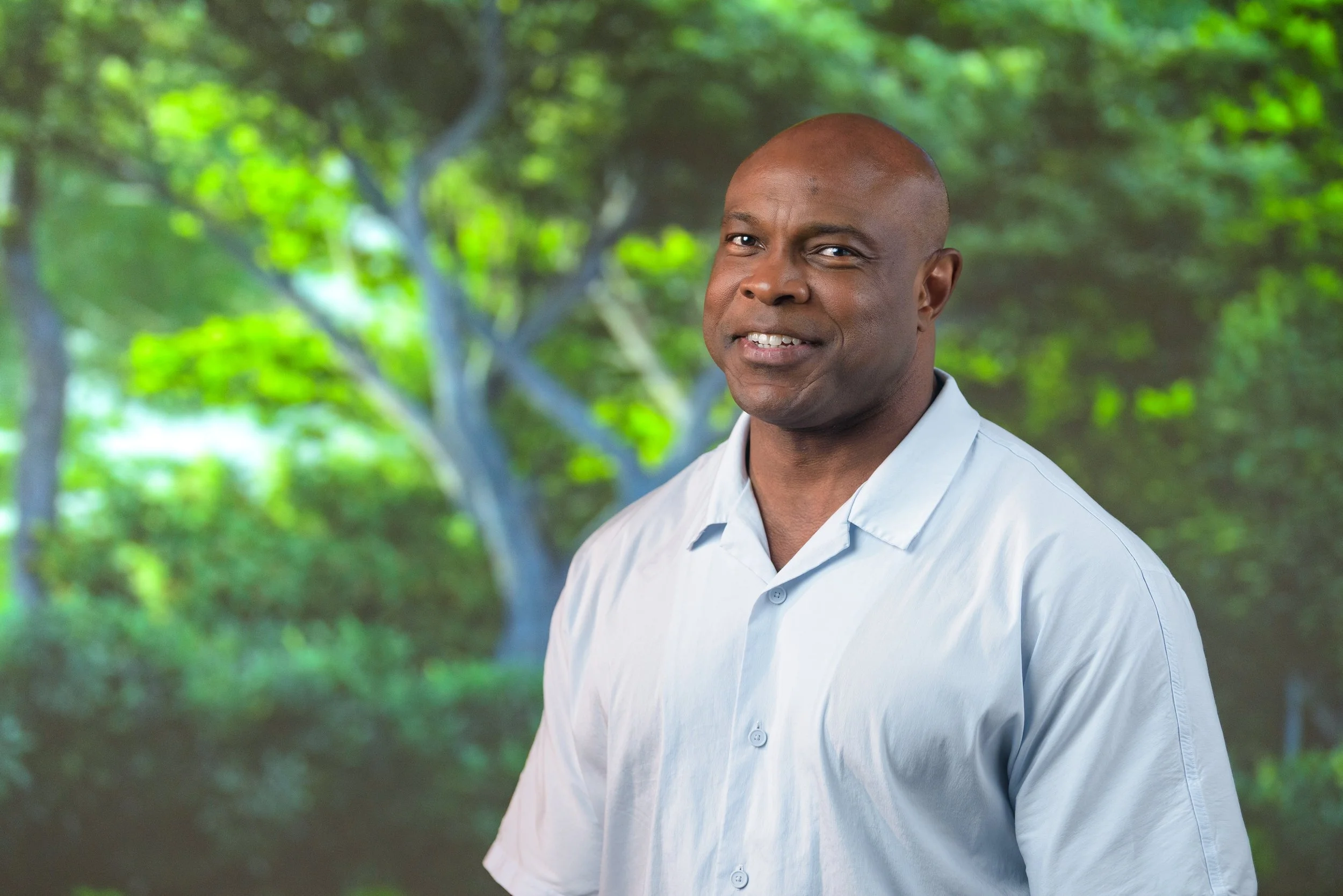 A man with a shaved head and a light blue button-up shirt standing outdoors in front of green trees and foliage.