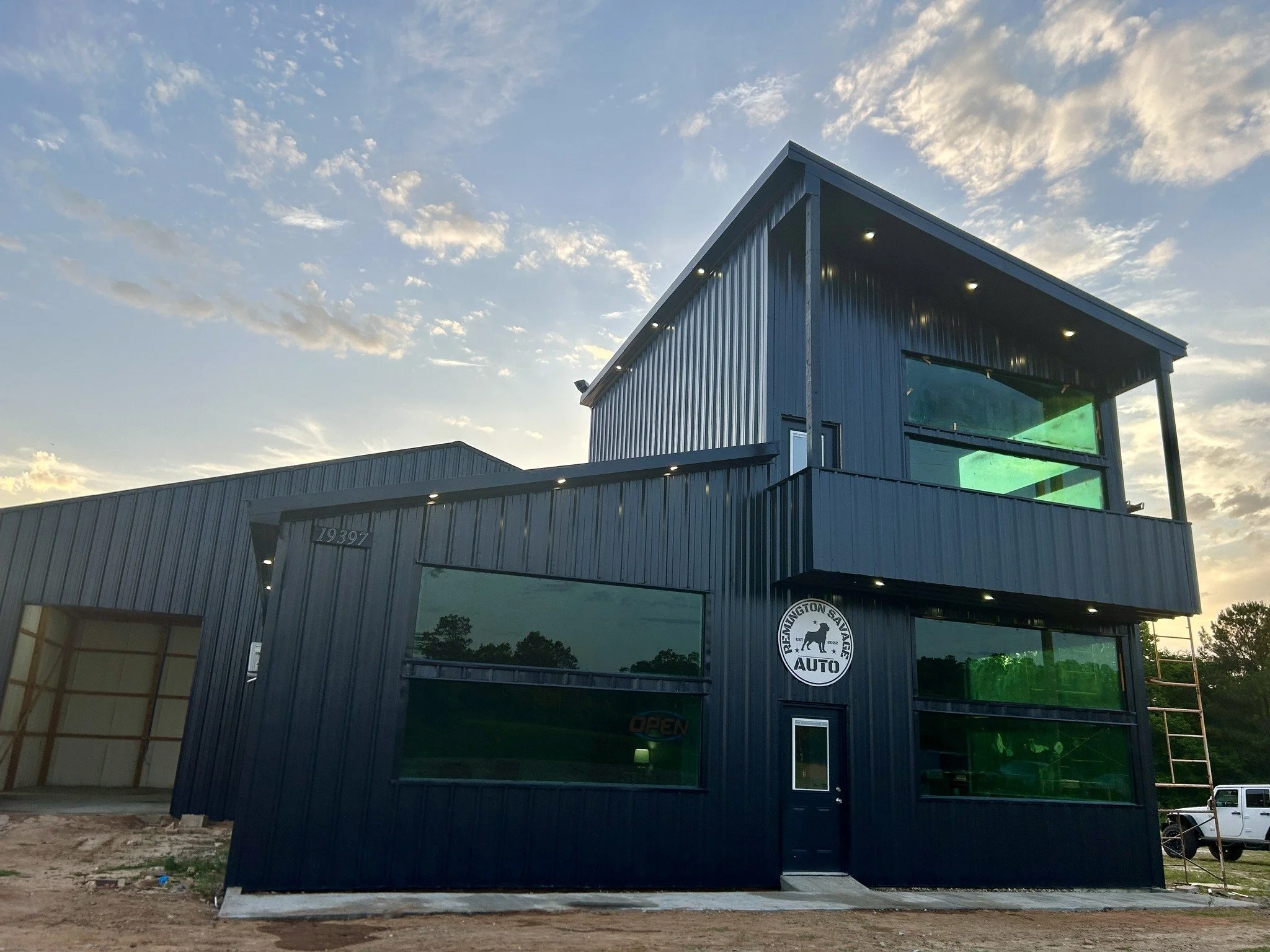 A modern, dark blue metal building with large windows, part of which has a green tint, and a circular logo that reads "Prescott Gunn Auto" with a silhouette of a dog. The building has exterior lighting and a sign indicating it is open. The sky above is partly cloudy with streaks of sunlight.