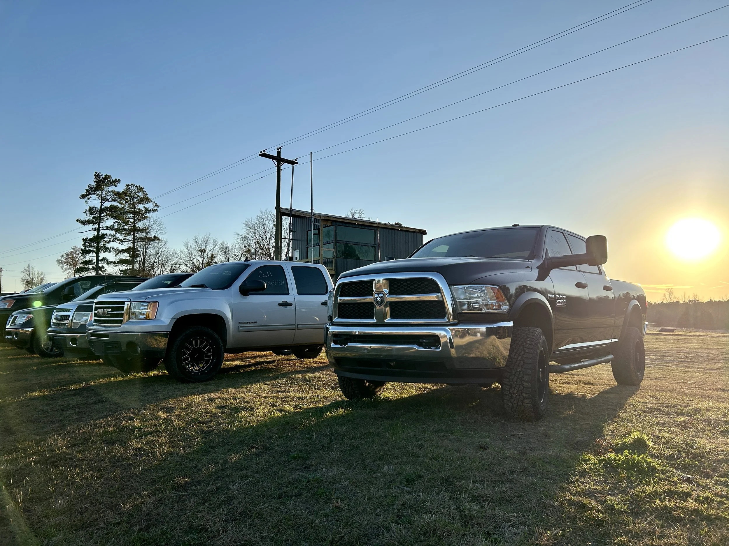 Row of parked trucks and cars on a grassy area during sunset with trees and power lines in the background.