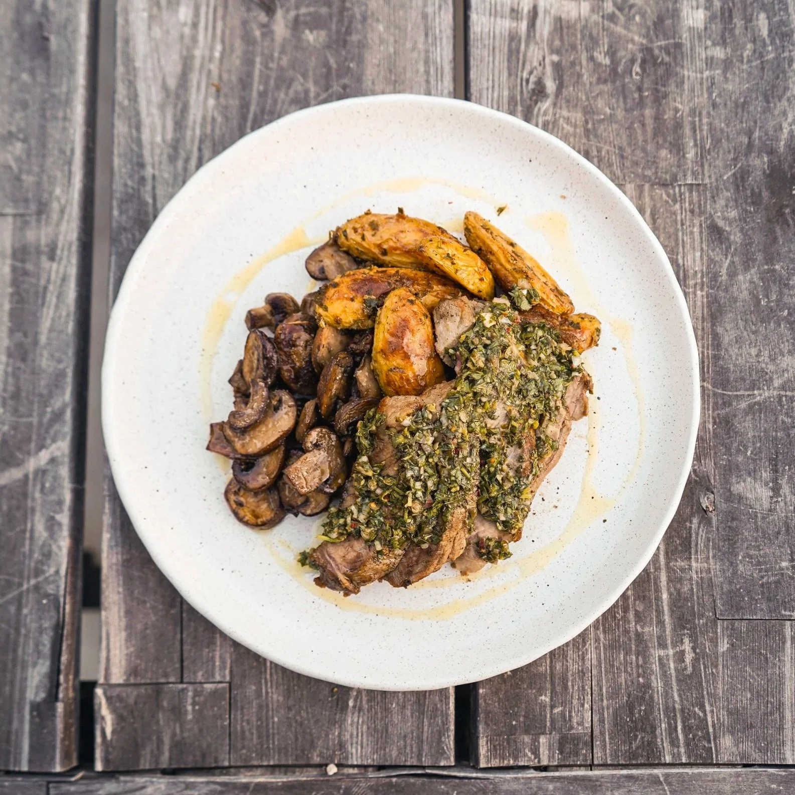 A white plate with roasted potatoes, sautéed mushrooms, and a piece of meat topped with chopped herbs on a rustic wooden table.