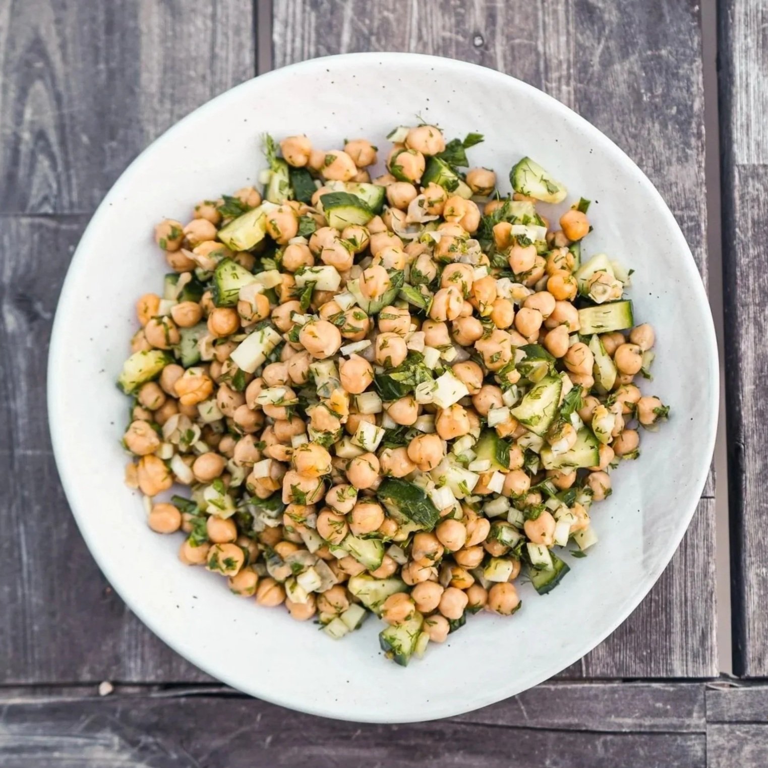 Plate of cooked chickpeas, diced cucumbers, and chopped herbs on a wooden surface.