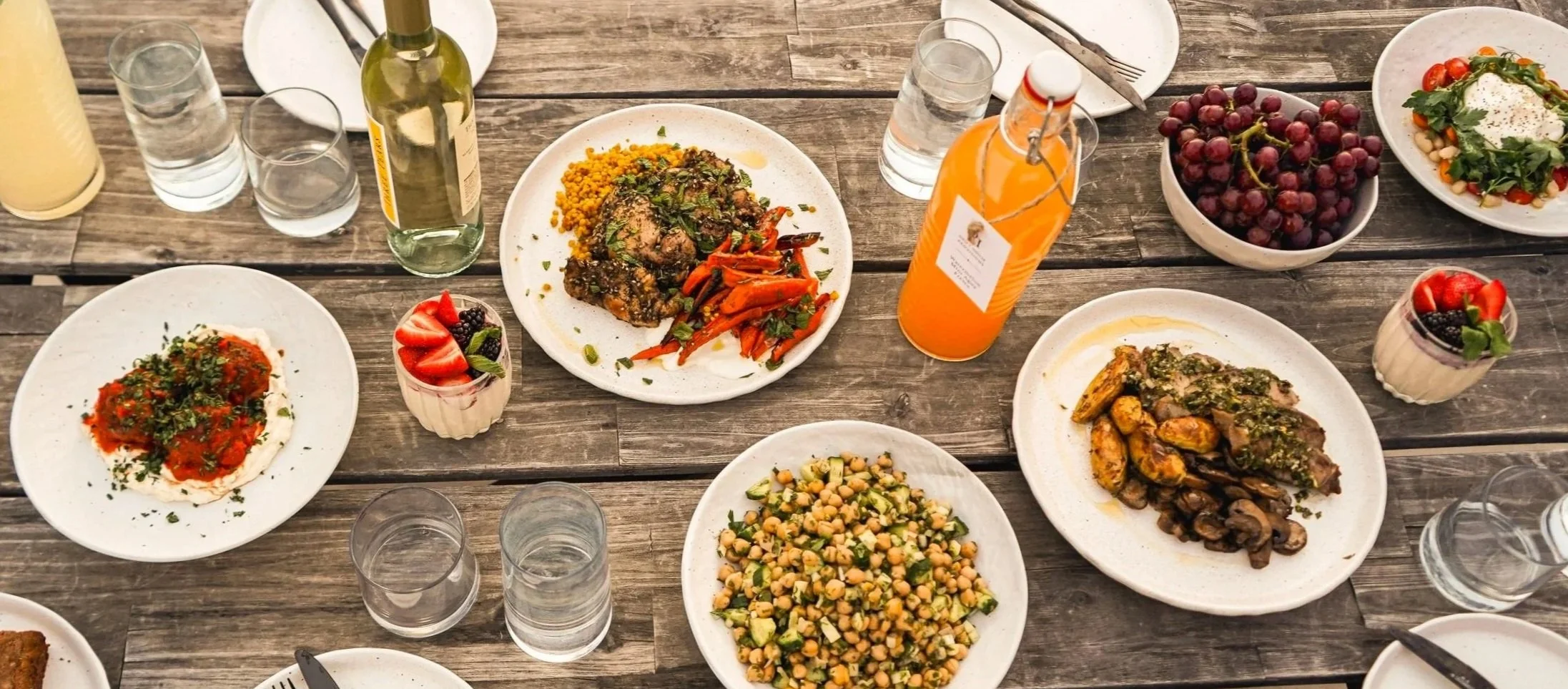 A variety of dishes on a wooden table including salads, roasted meats, chickpea salad, a yogurt dessert with berries, and bottles of orange juice and wine.
