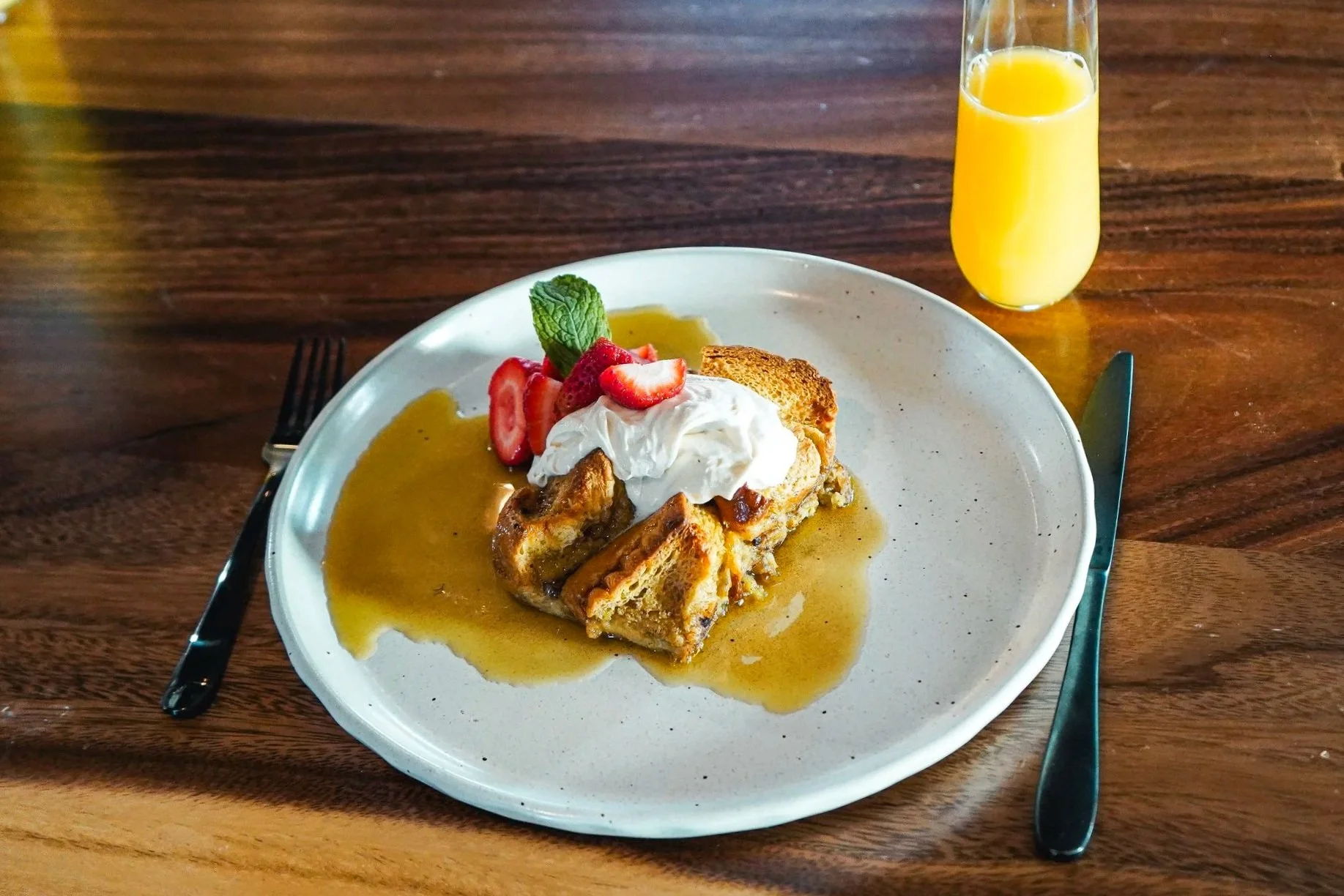 A plate of French toast topped with whipped cream and strawberries, served with syrup, accompanied by a glass of orange juice, on a wooden table.