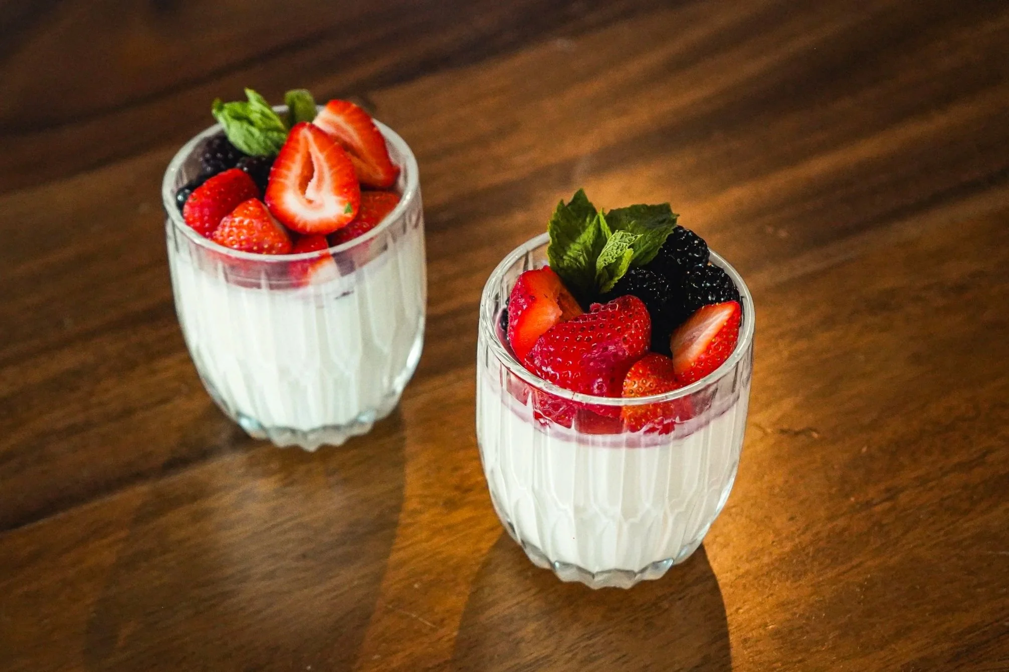 Two glasses of yogurt topped with strawberries, blackberries, and mint leaves on a wooden table.