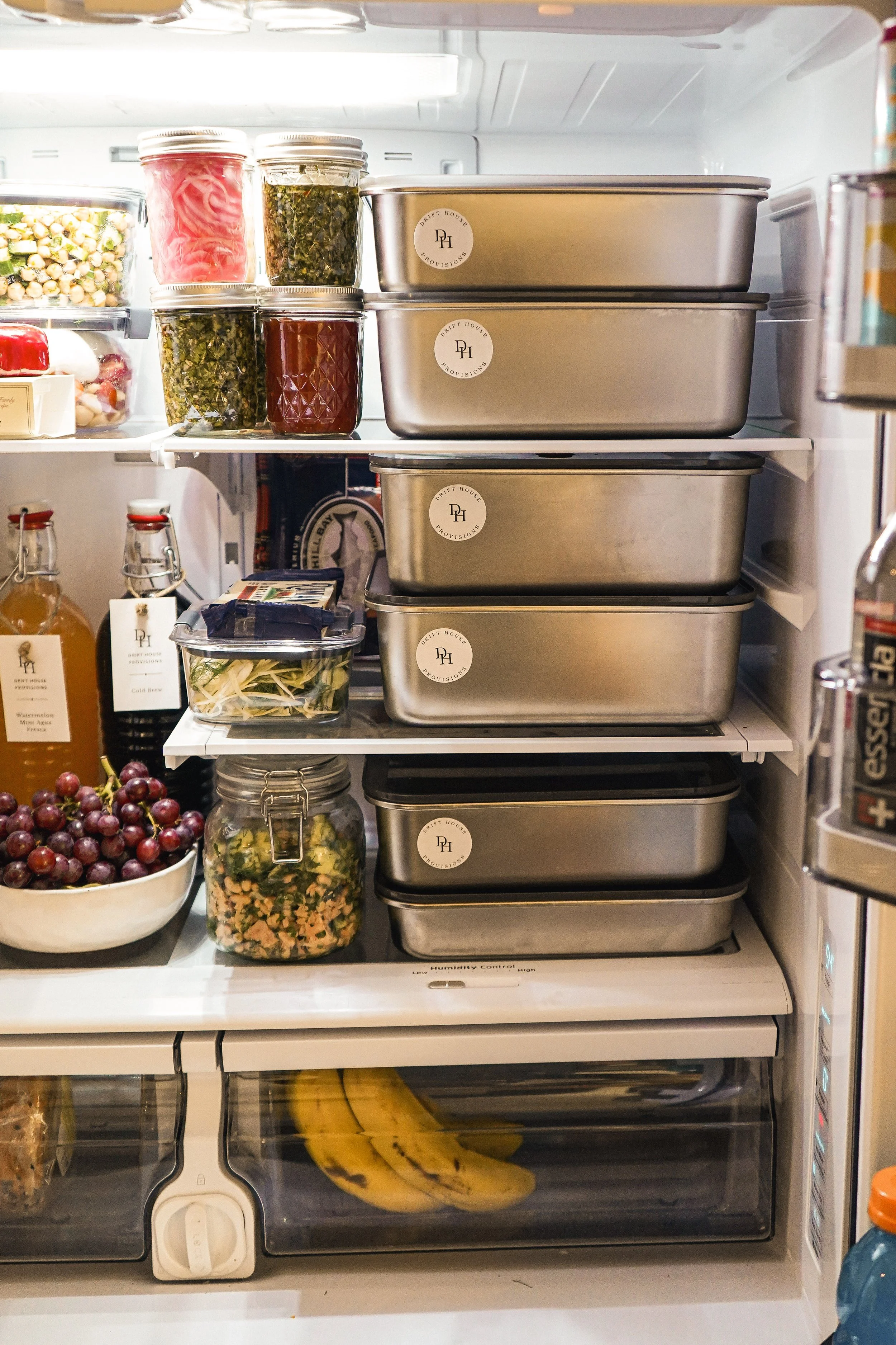 Inside a refrigerator, glass jars contain vegetables, fruits, and sauces; plastic containers hold prepared foods; bananas are on a lower shelf; grapes are in a bowl; and a can of soda is on the right.