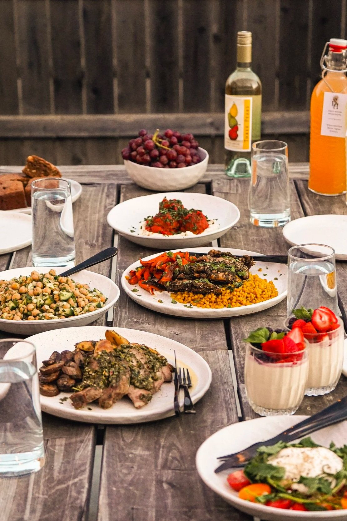 Dinner table with various dishes, including grilled meat, chickpea salad, roasted vegetables, grapes, strawberries, and drinks.