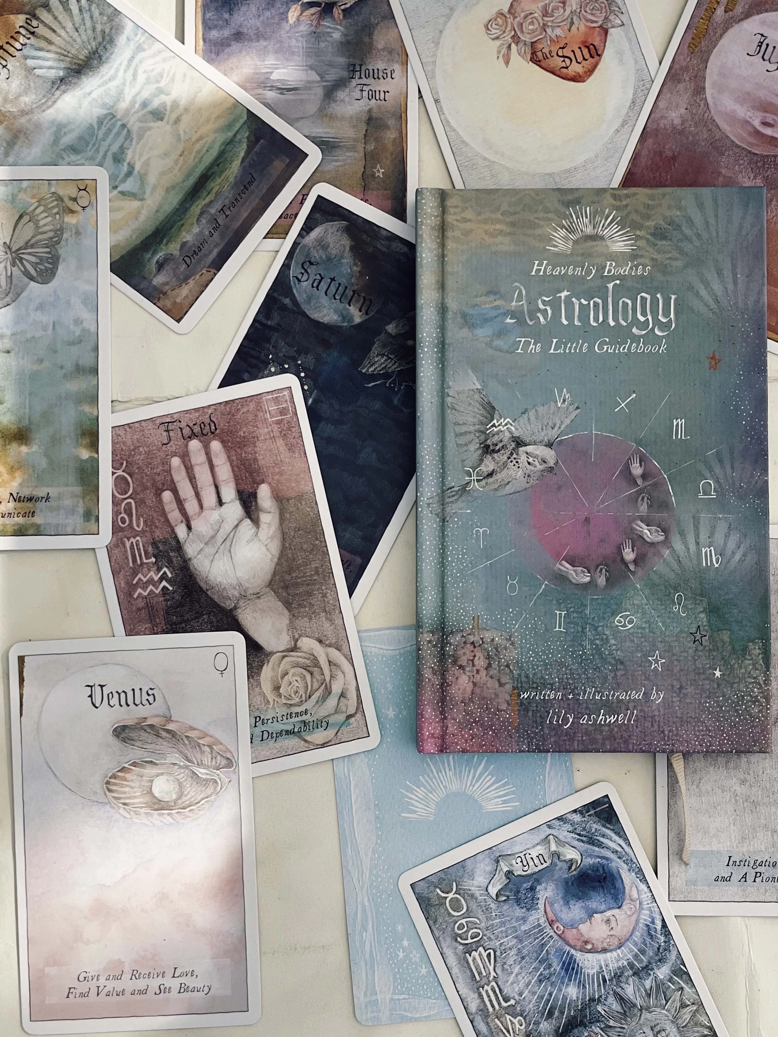 A book titled 'Heavenly Bodies Astrology The Little Guidebook' by Lily Ashwell surrounded by various astrology-themed tarot cards featuring symbols and illustrations of moons, stars, and astrological signs.