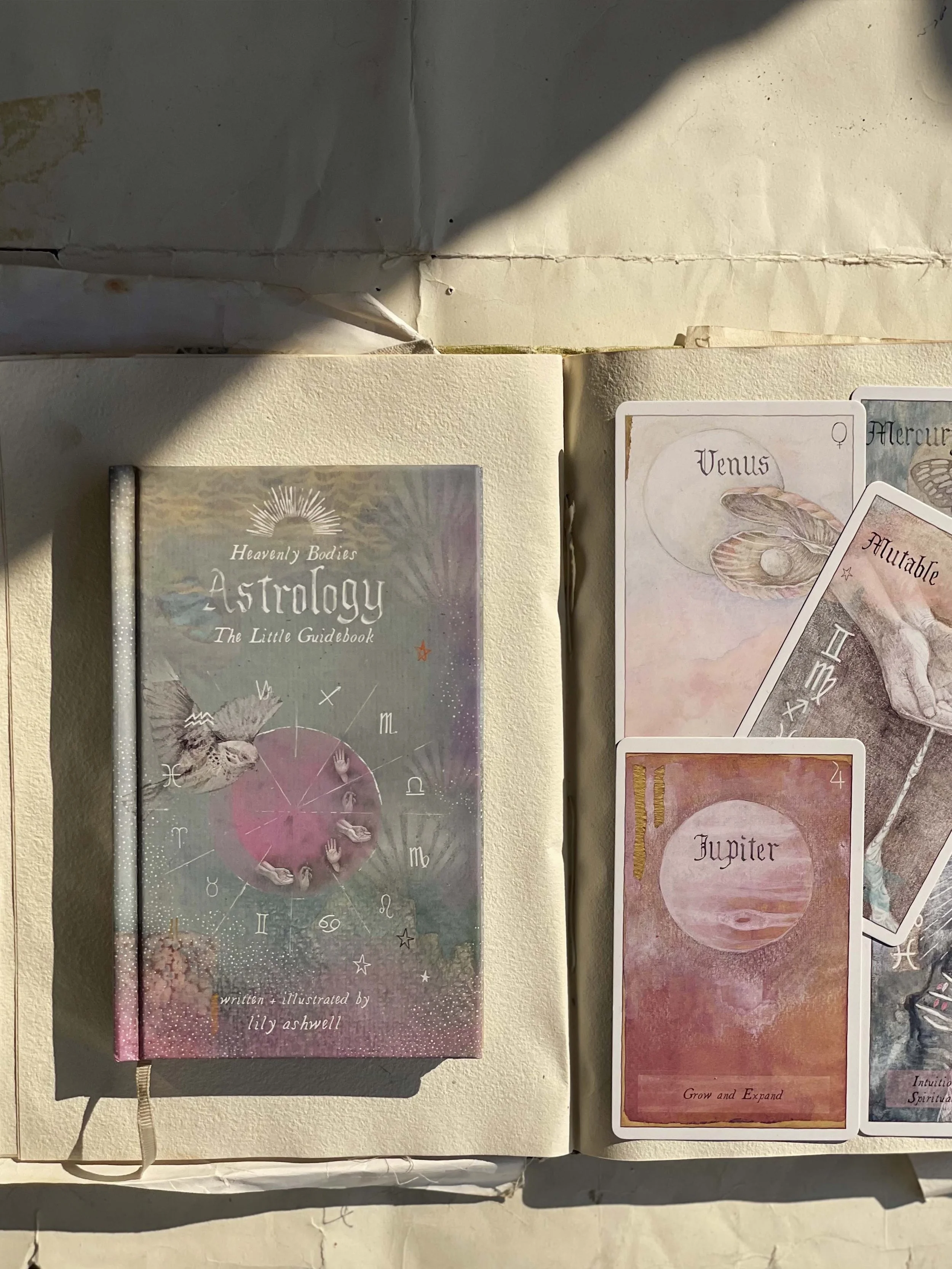 A book titled 'Heavenly Bodies Astrology The Little Guidebook' by Lily Ashwell along with several astrology-themed tarot cards, including Venus, Jupiter, and a mutable card, displayed on a light-colored surface with sunlight and shadows.