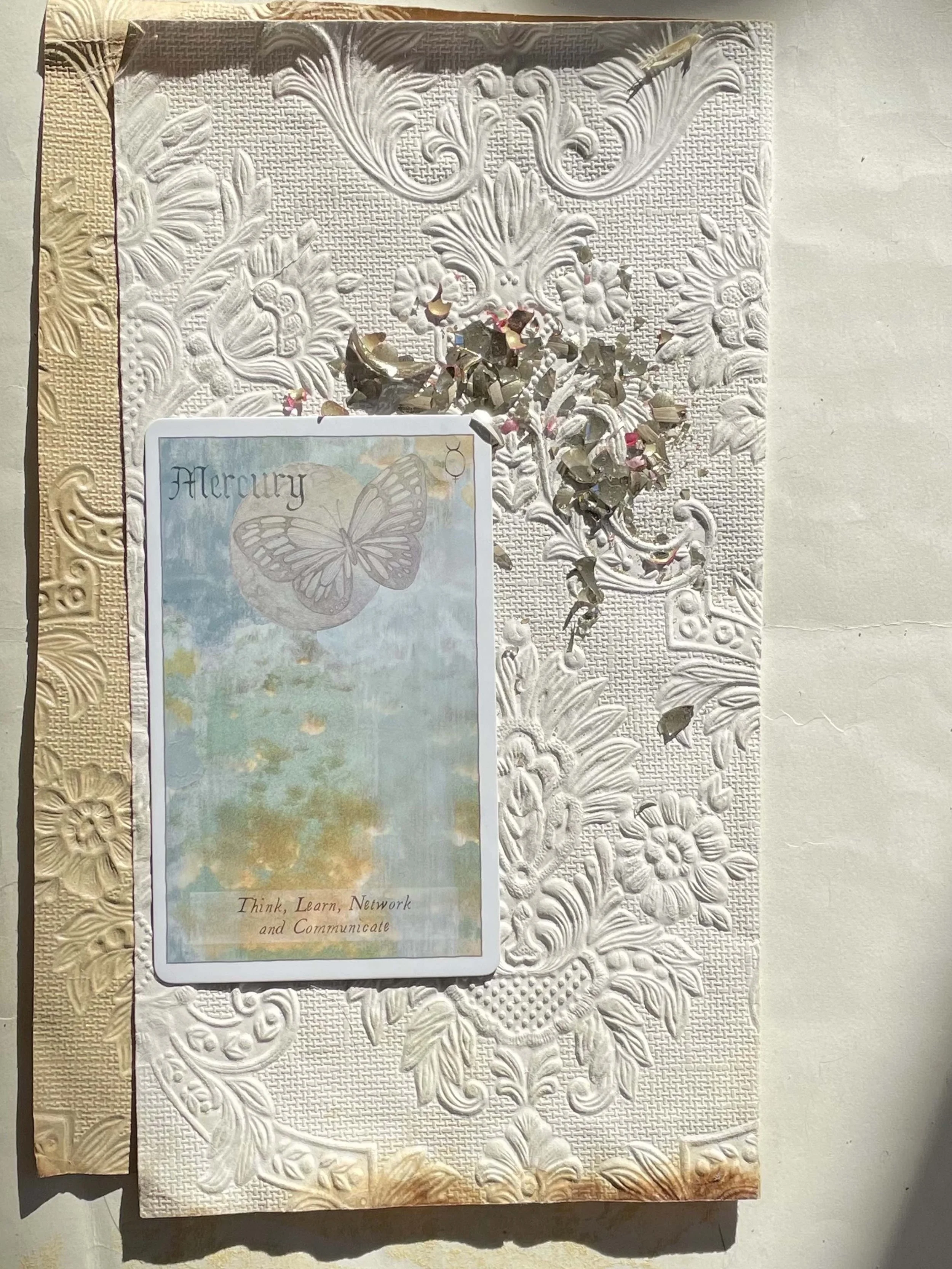 Decorative white embossed surface with metallic confetti, a card from Lily Ashwell's Heavenly Bodies Astrology Deck featuring a butterfly and the word 'Mercury', and gold decorative tape.