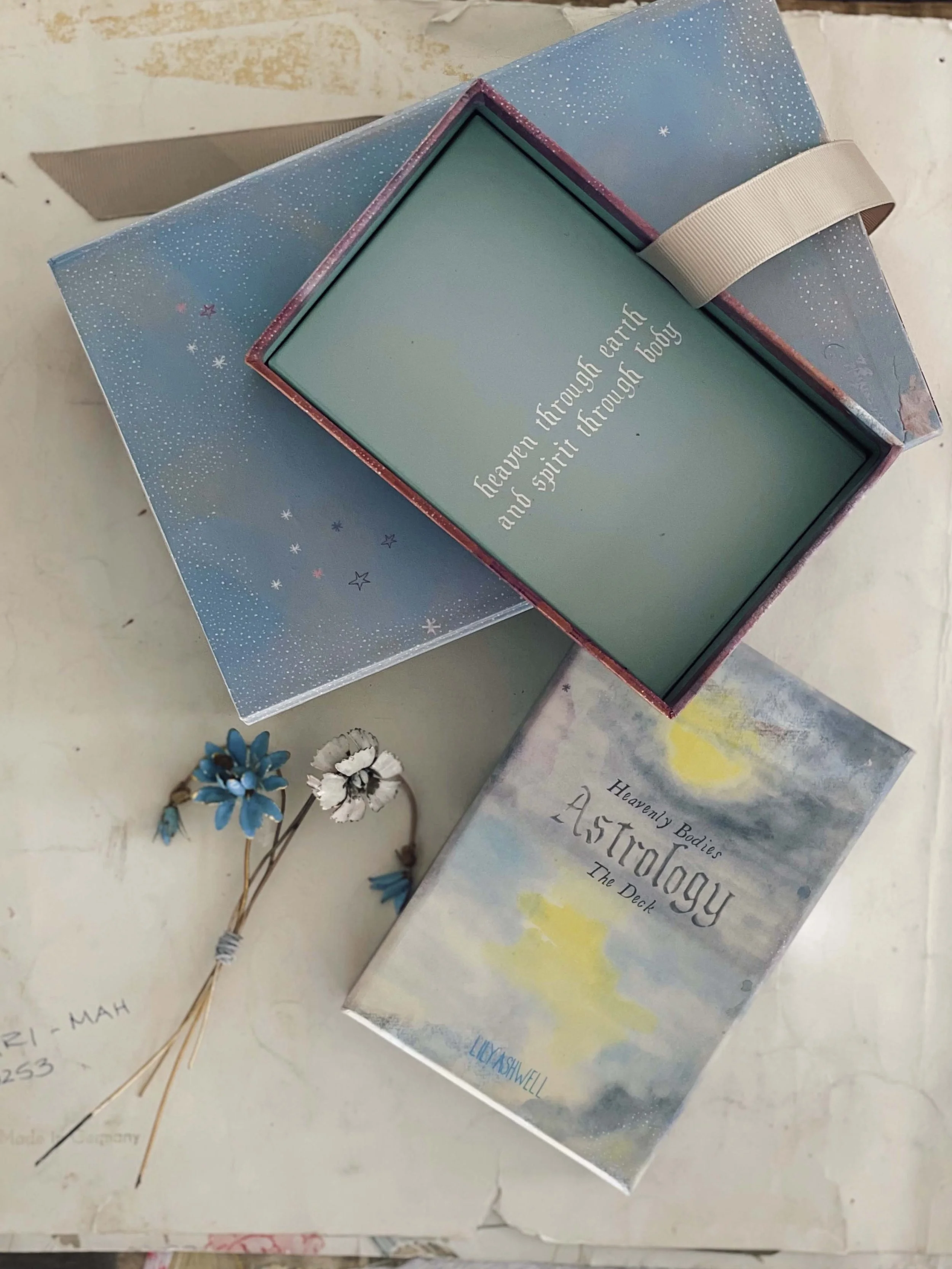 A decorated box with a stars and snowflake pattern, a book titled 'Heavenly Bodies Astrology The Deck', a small bouquet of blue and white flowers, and a framed quote reading 'heaven through earth and spirit through body'.