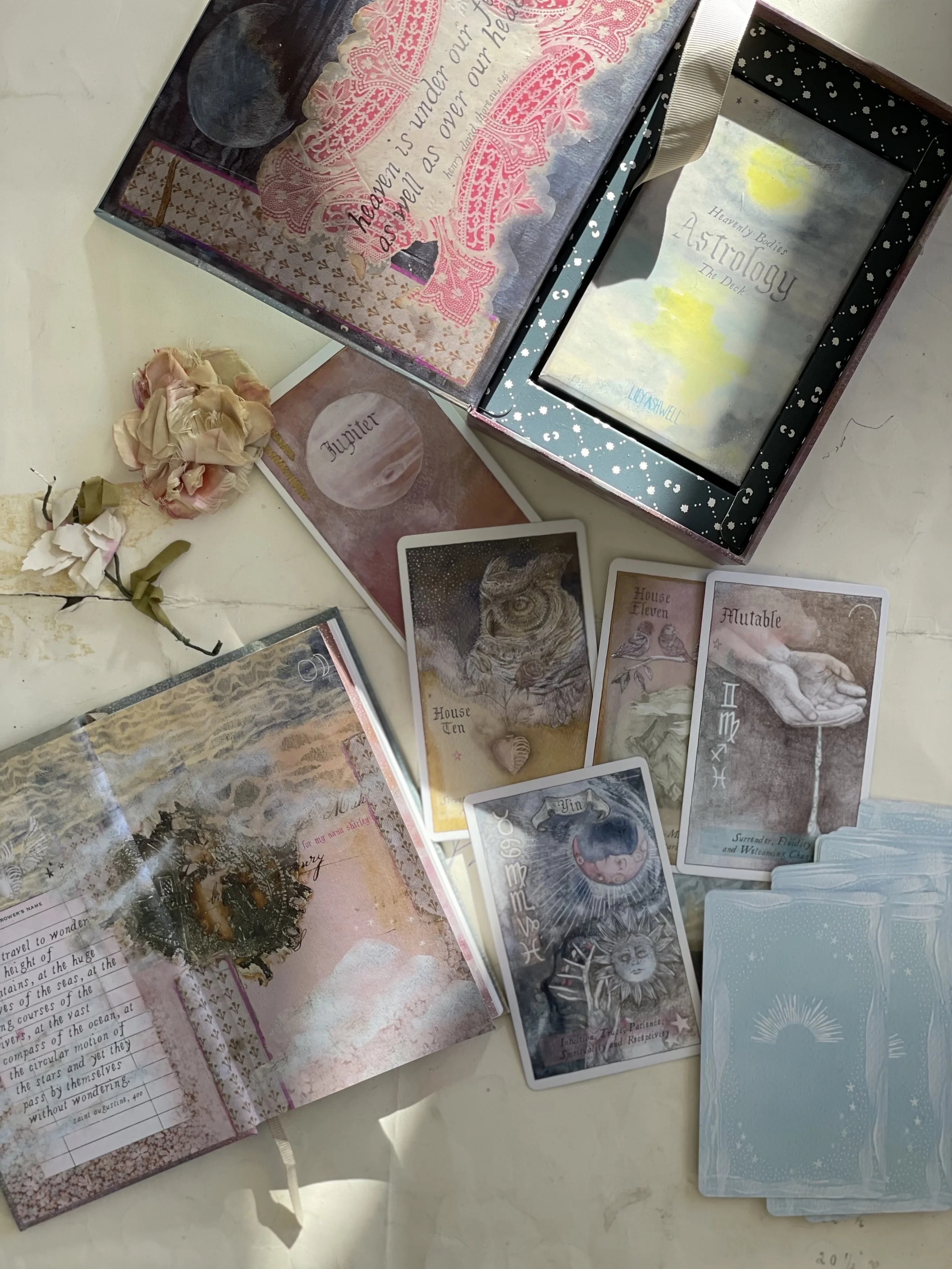A collection of oracle cards, from the Heavenly Bodies Astrology Deck – an open decorative book, a small pink flower, and a box with themed cards on a white surface.