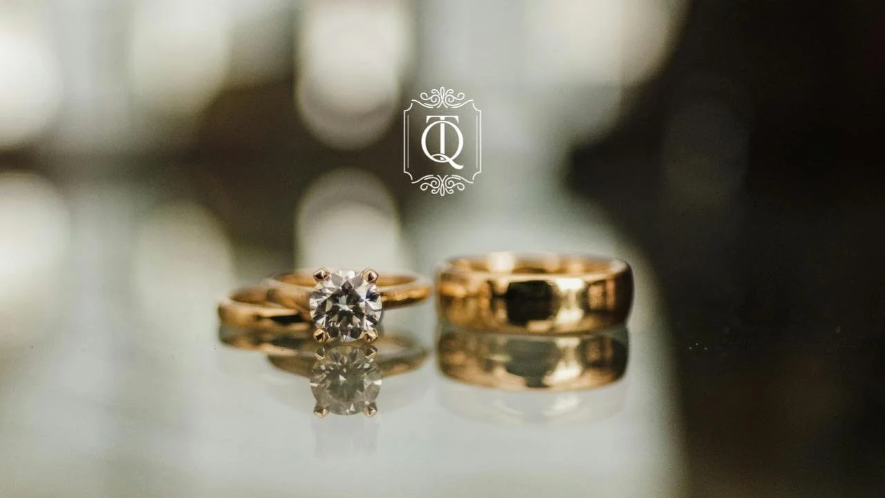 Dois anéis de ouro, um com um diamante central e outro mais simples. Ambos estão refletidos sobre uma superfície.