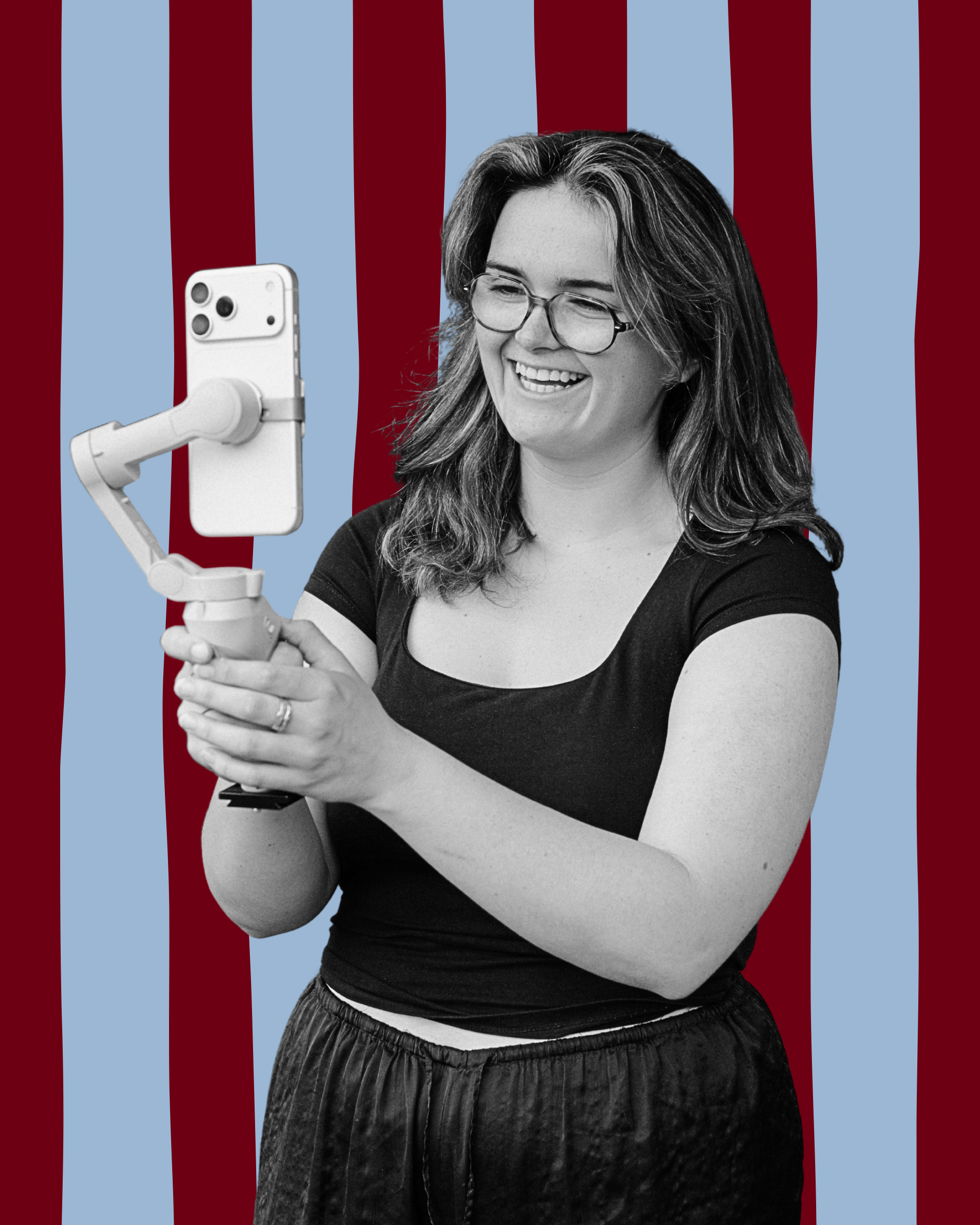 A smiling woman with glasses, wearing a black T-shirt, holding a smartphone mounted on a handheld stabilizer, taking a selfie against a background with vertical red and light blue stripes.