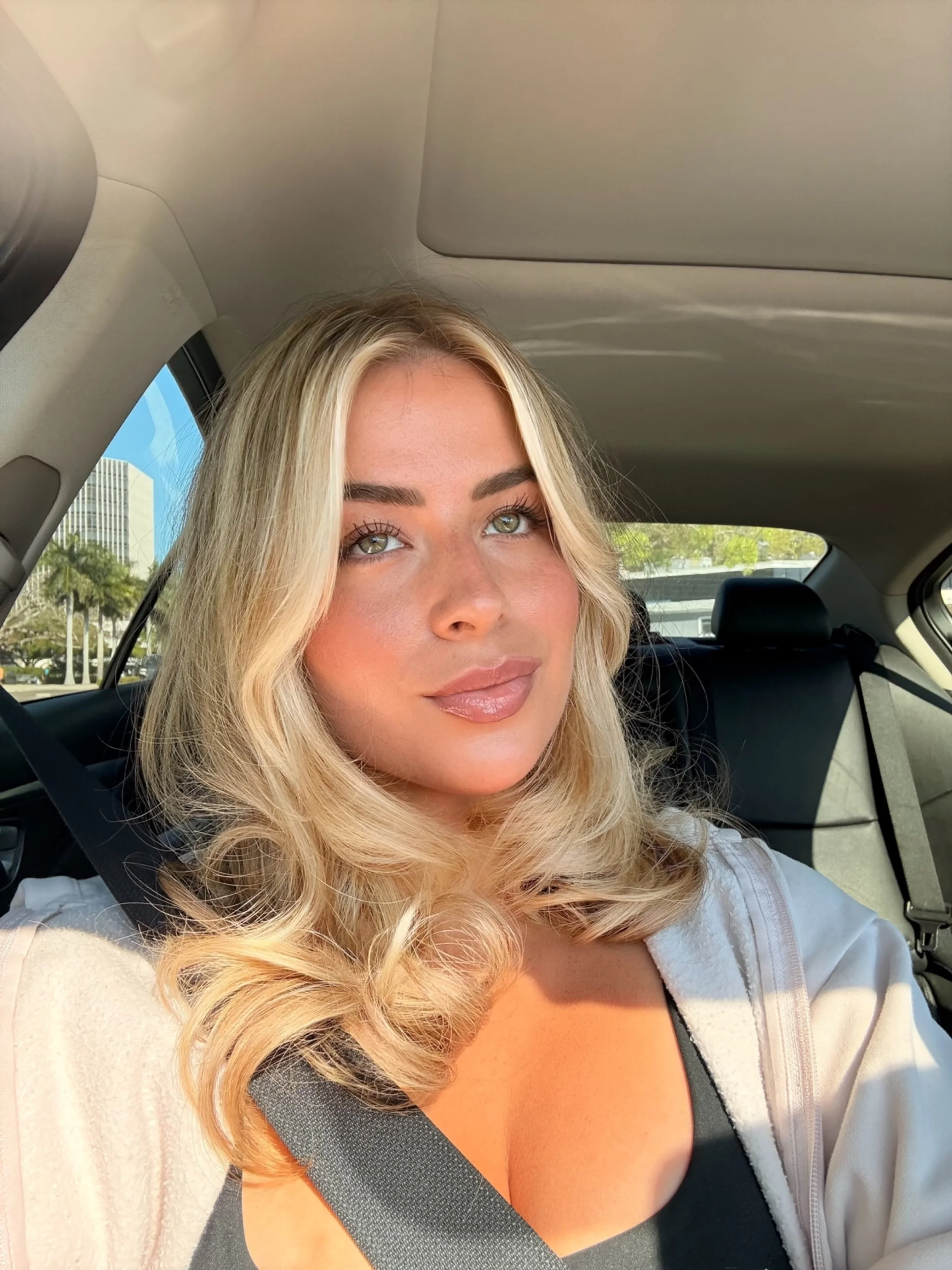 A woman with blonde hair and green eyes sitting in the backseat of a car, wearing a black top and a white zip-up jacket, with a seatbelt across her chest. The background shows a cityscape with a tall building and trees.
