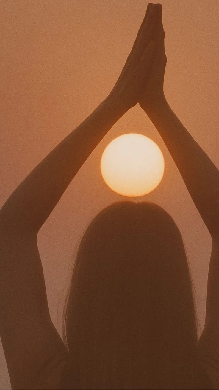 A person with long hair has their arms raised above their head, with palms pressed together, creating a silhouette against the setting or rising sun.