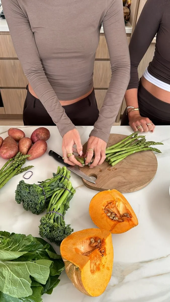 Person peeling asparagus on a round wooden cutting board in a kitchen, with potatoes, broccoli, romaine lettuce, and a halved pumpkin on the white marble countertop.