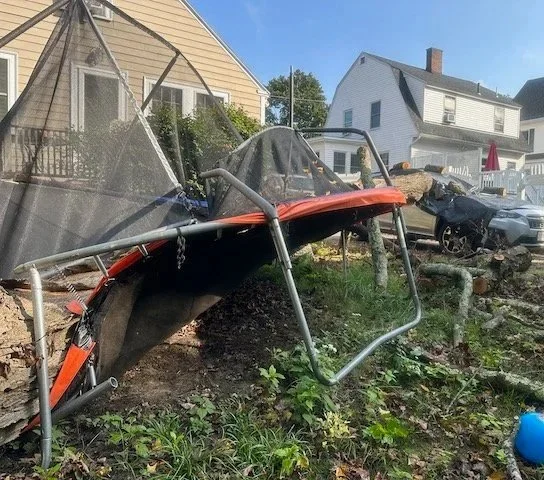 A toppled trampoline with a broken safety net in a yard, with damaged cars and house in the background.